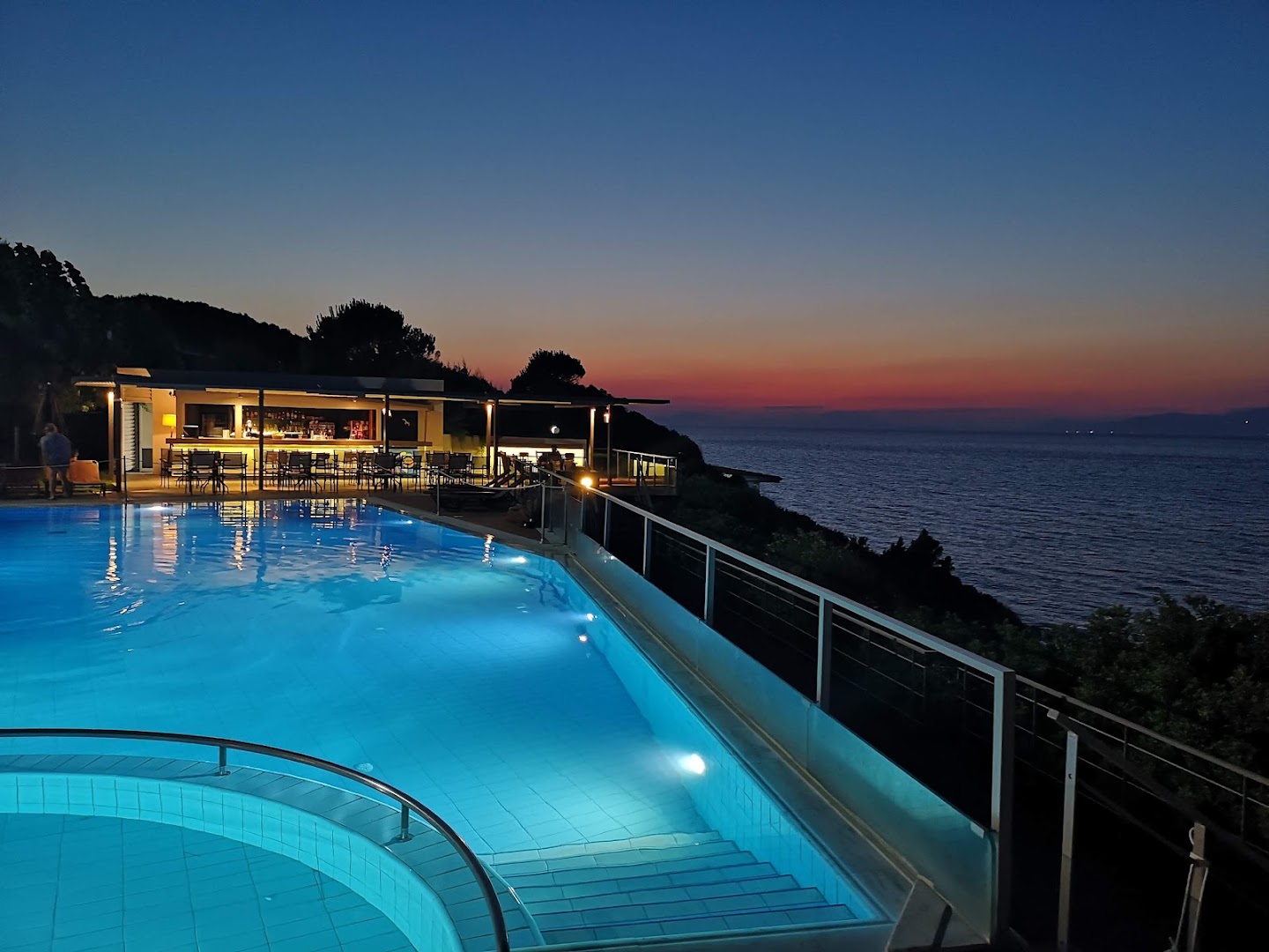 Niforeika Beach Hotel izgaismots baseins un bārs vēlā vakarā uz jūras fona — romantiska atmosfēra un saulrieta atblāzma