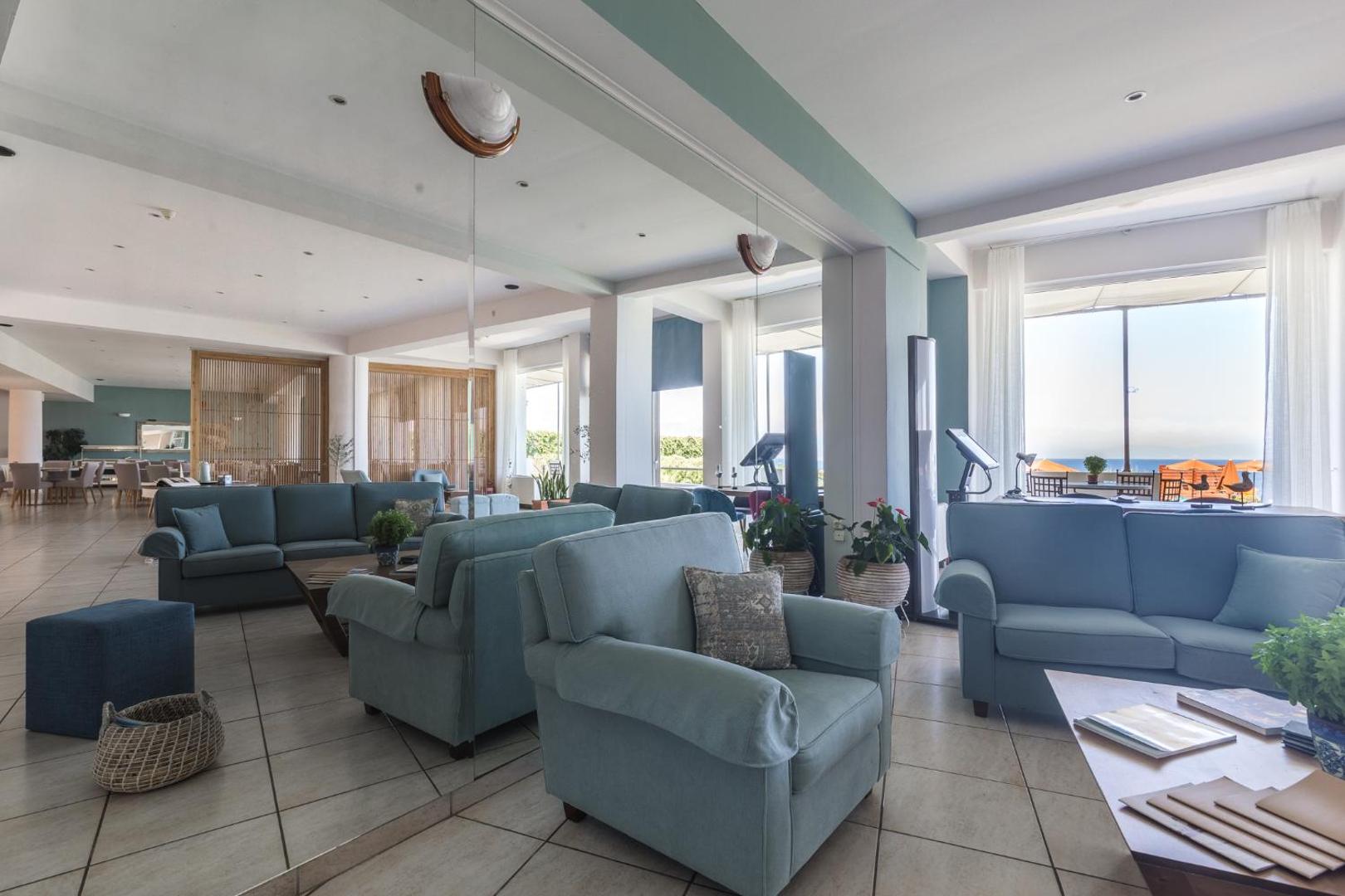 Niforeika Beach Hotel lobby atpūtas telpa ar gaiši ziliem dīvāniem un lieliem logiem, pa kuriem paveras skats uz jūru