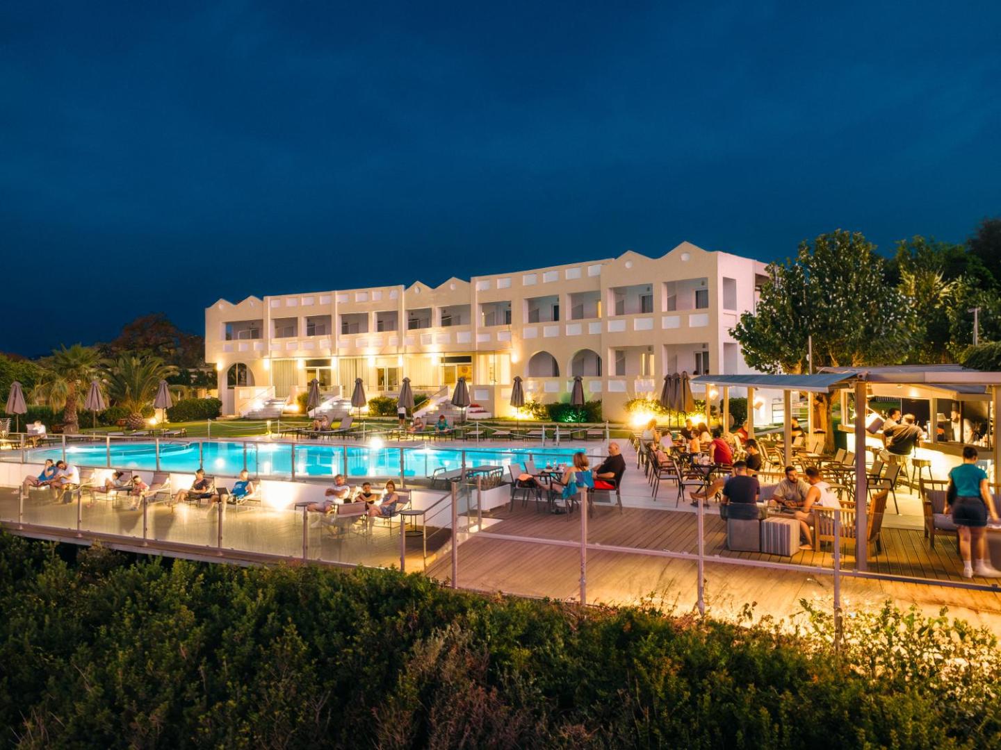 Niforeika Beach Hotel galvenā ēka un baseina zona vakarā ar apgaismojumu — viesi bauda vakariņas un dzērienus pie baseina bāra