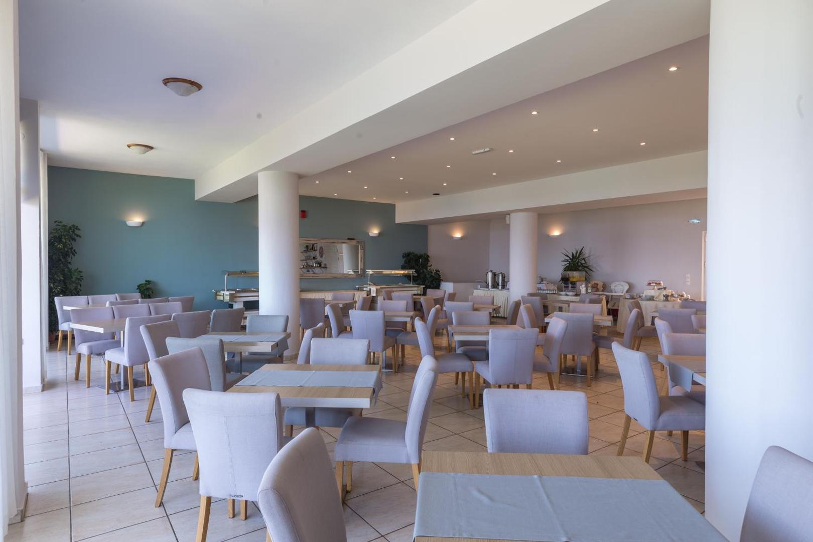 Niforeika Beach Hotel galvenais restorāns ar gaišām mēbelēm, kolonnām un bufetes zonu — plaša un saulaina telpa brokastīm