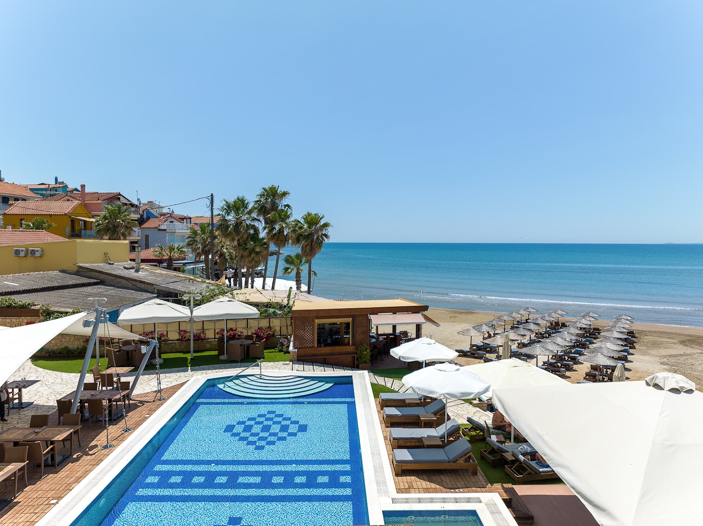 Almira Hotel Arkoudi skats uz baseinu un privāto pludmali Grieķijā — piekrastes panorāma ar saulessargiem, palmām un zilu jūru