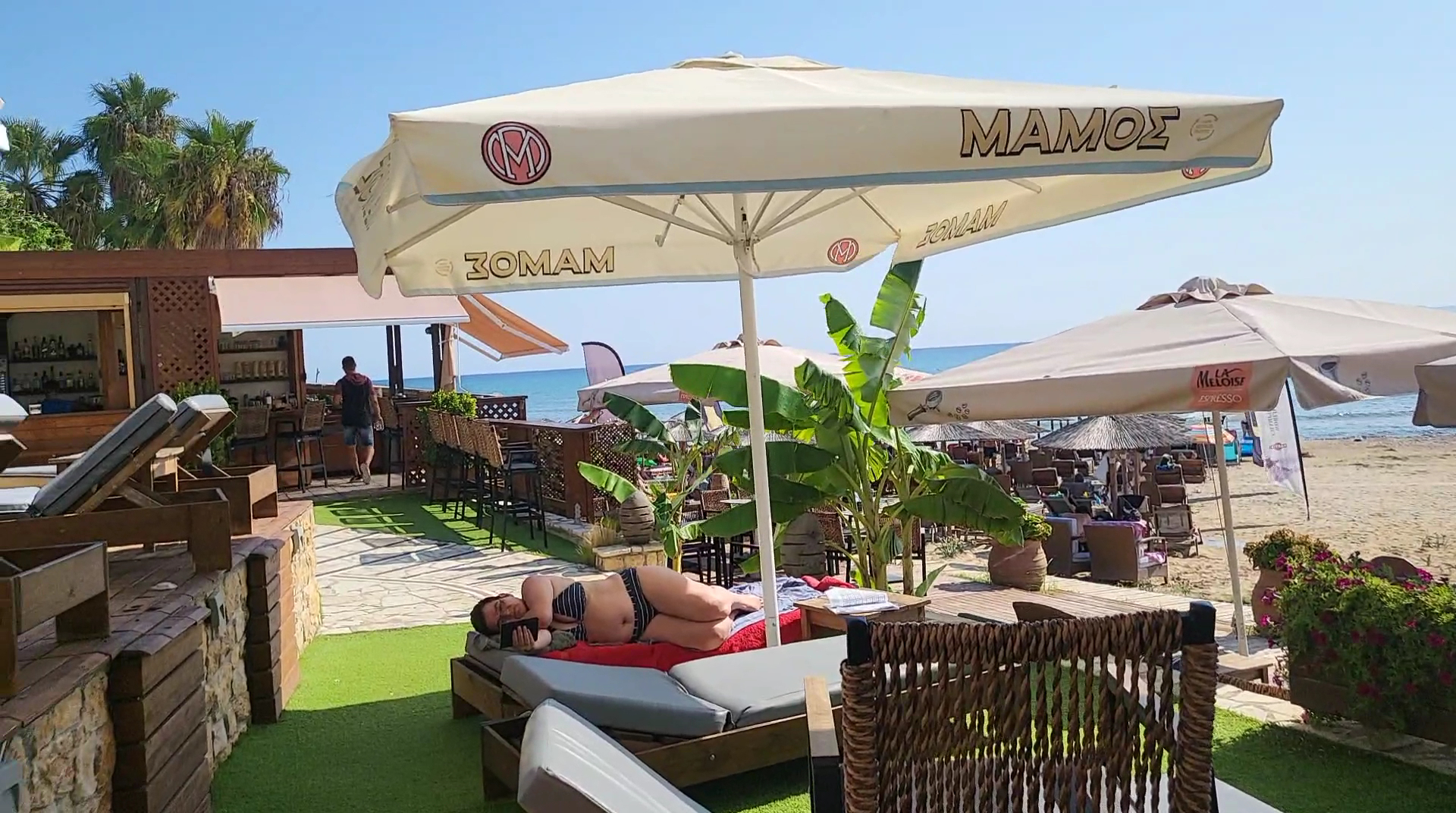 Almira Hotel pludmales bārs ar atpūtas krēsliem un saulessargiem — viesi relaksējas piekrastē netālu no bāra zonas