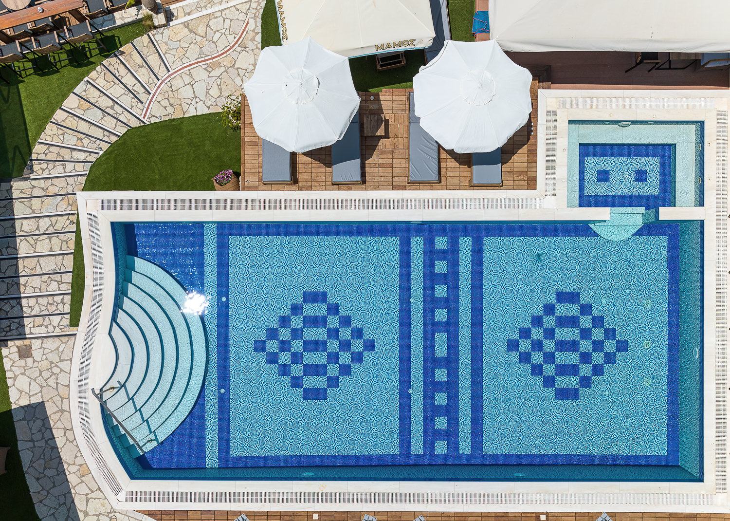 Almira Hotel Arkoudi āra baseins no putna lidojuma skata — zilas mozaīkas flīzes, sauļošanās krēsli un saulessargi uz koka terases