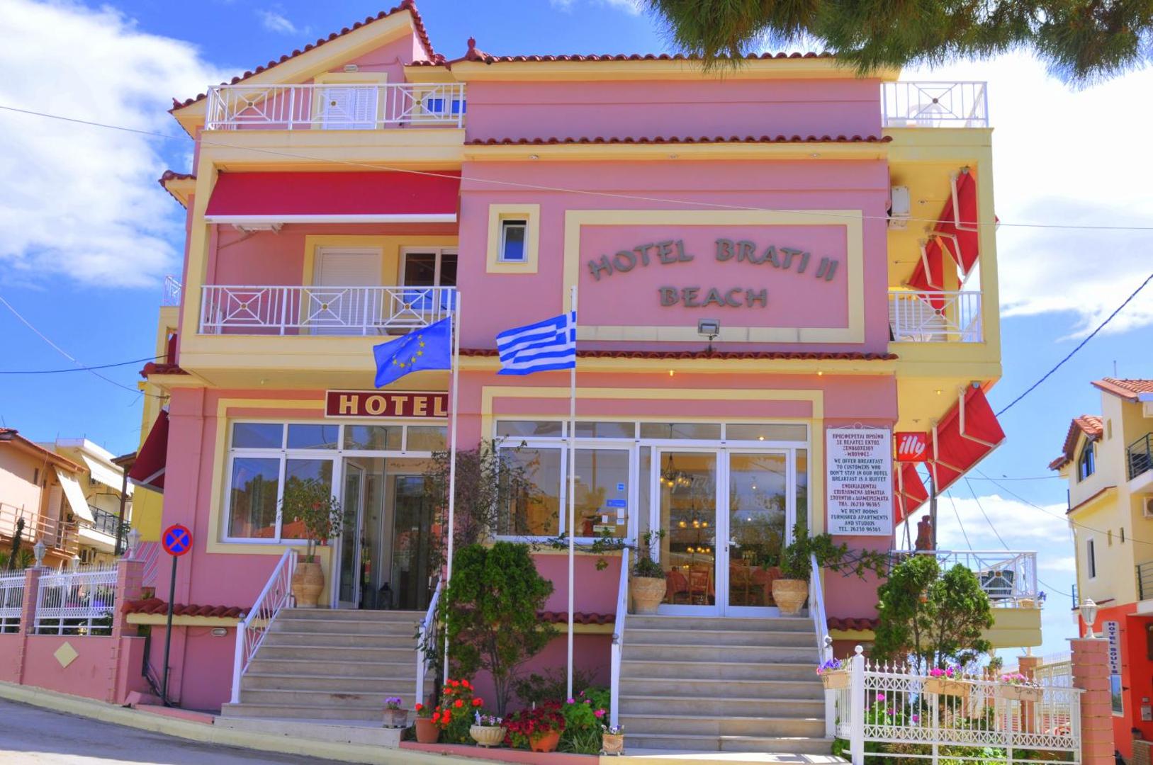 Brati II Beach Hotel rozā fasāde Grieķijā — trīs stāvu ēka ar balkoniem, karogiem un ieeju piekrastes pilsētā