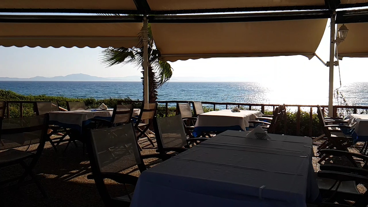 Brati II Beach Hotel āra terases restorāns ar skatu uz Jonijas jūru saulrietā, balti klāti galdi zem nojumes.
