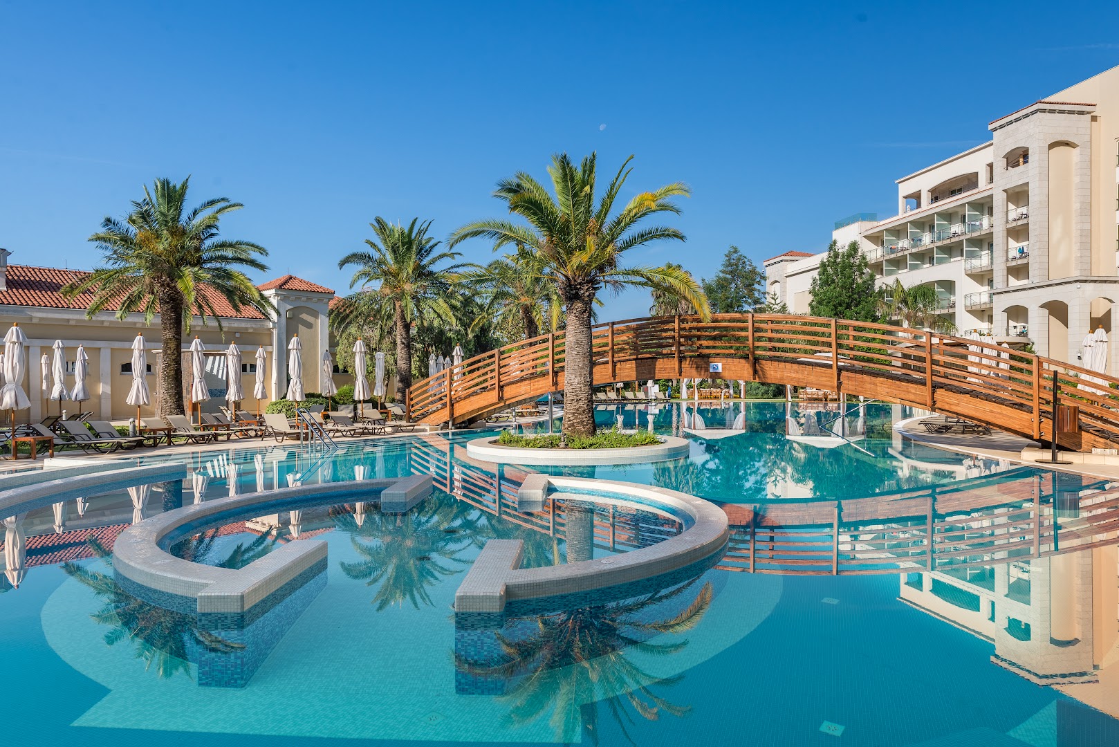 Splendid Conference & Spa Resort āra baseins ar koka tiltiņu un palmām Bečiči kūrortā, Melnkalnē — atpūtas zona ar saulessargiem
