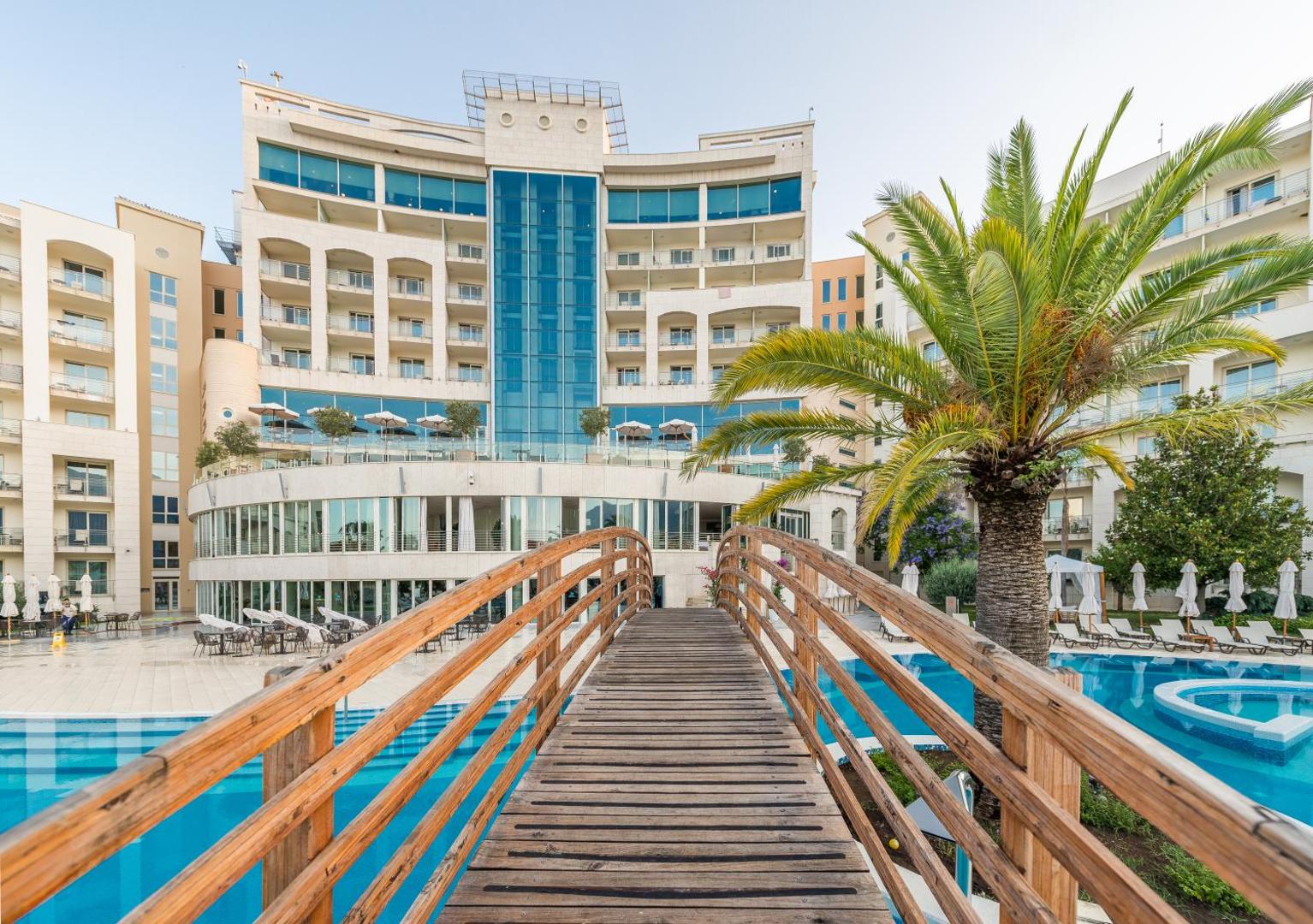 Koka tiltiņš pār zilu āra baseinu ar skatu uz Splendid Conference & Spa Resort galveno ēku un palmām