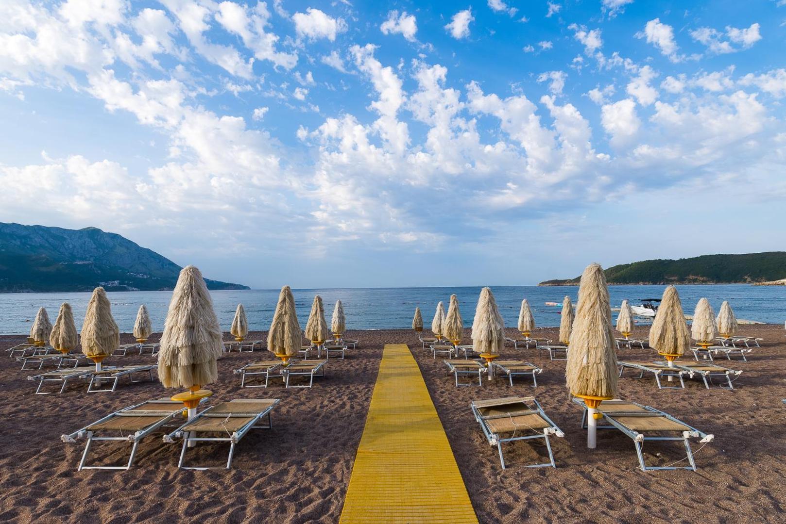 Skats no Splendid Conference & Spa Resort pludmales uz jūru un kalniem — dzeltena koka laipa starp saulessargiem zem mākoņainām debesīm