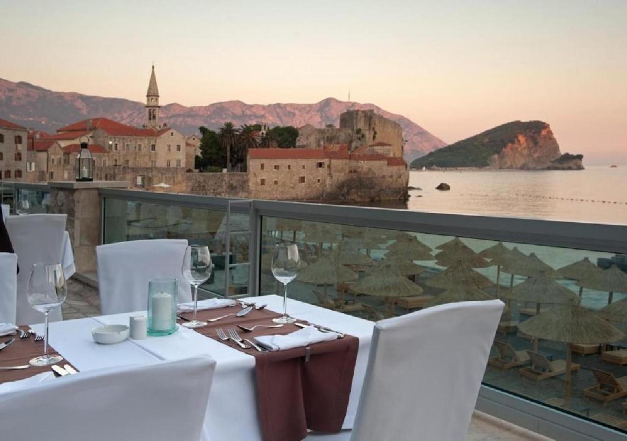 Avala Resort & Villas restorāna terase ar skatu uz Budvas vecpilsētu un Adrijas jūru saulrietā — eleganti klāti galdi