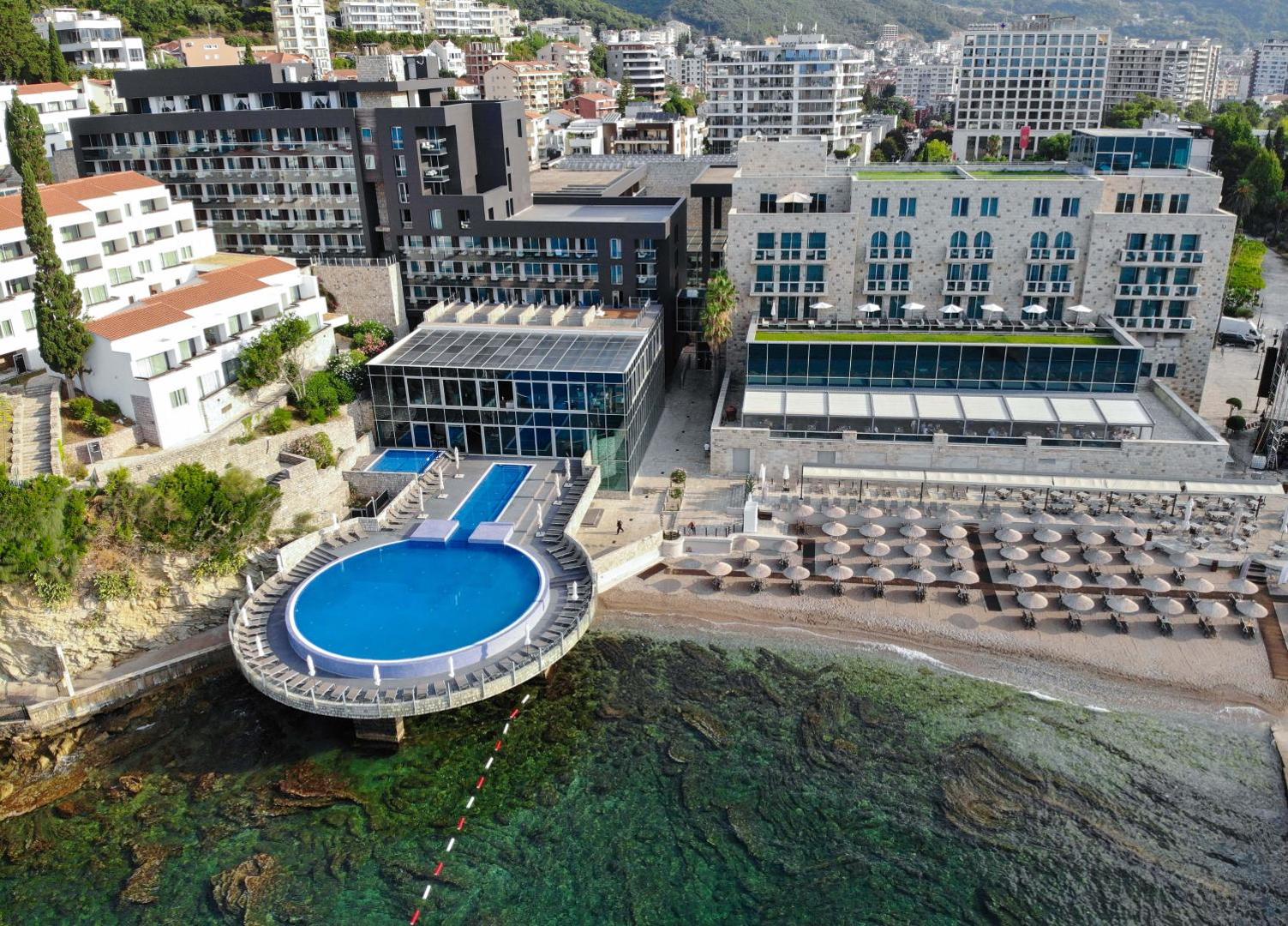 Avala Resort & Villas skats no putna lidojuma Budvā, Melnkalnē — unikāls apaļš baseins virs jūras, viesnīcas ēkas un privātā pludmale