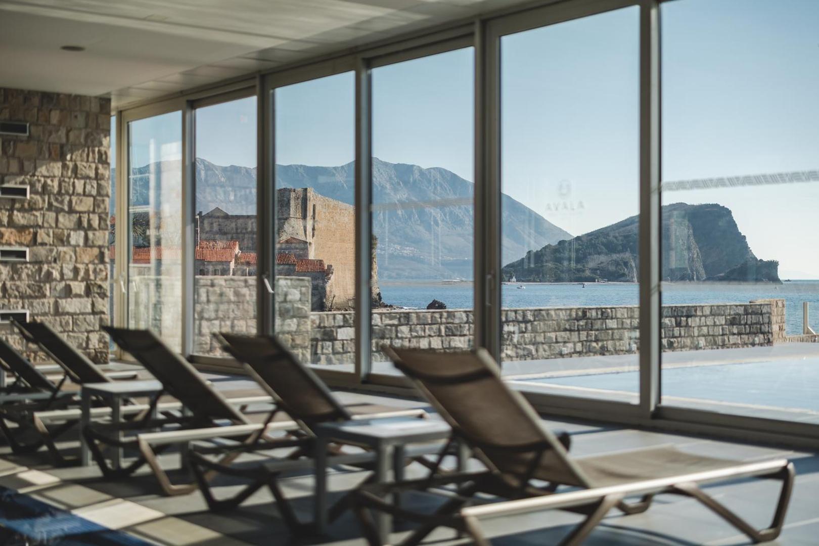 Skats no Avala Resort & Villas iekštelpu atpūtas zonas caur lieliem logiem uz Budvas vecpilsētu un kalniem