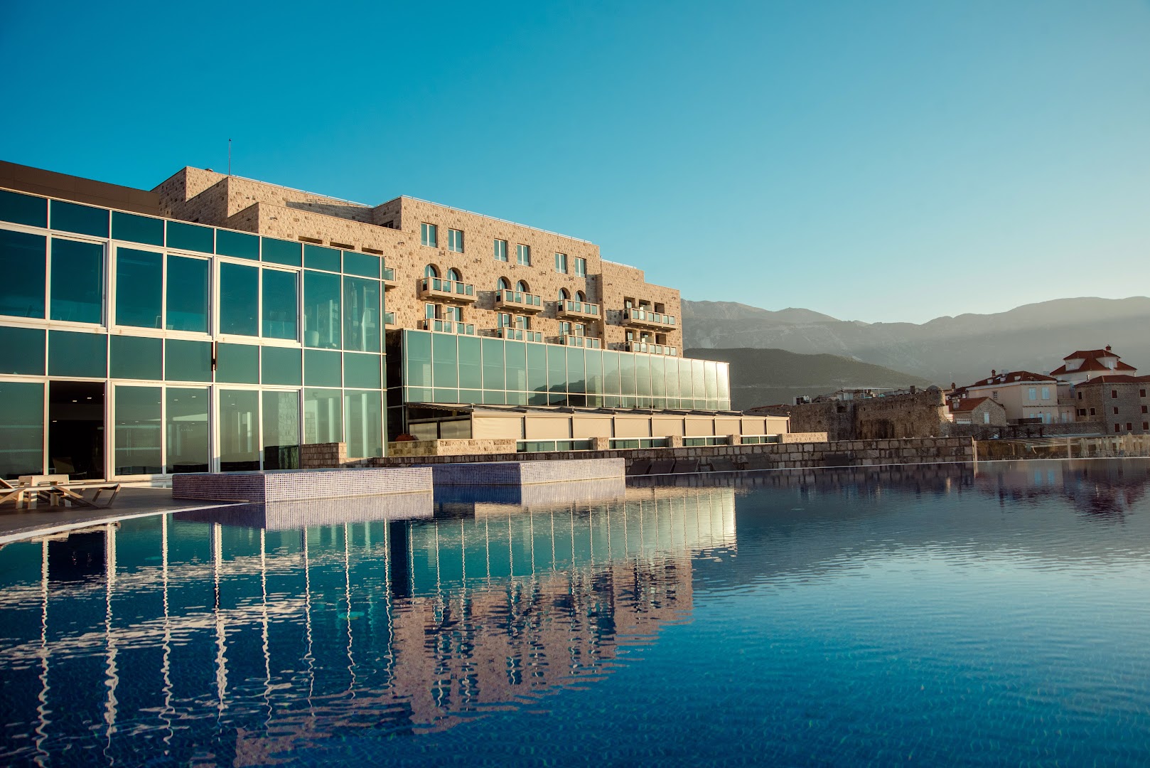 Avala Resort & Villas modernā arhitektūra Budvā ar lielu āra baseinu, kurā atspīd viesnīcas fasāde un apkārtējie kalni