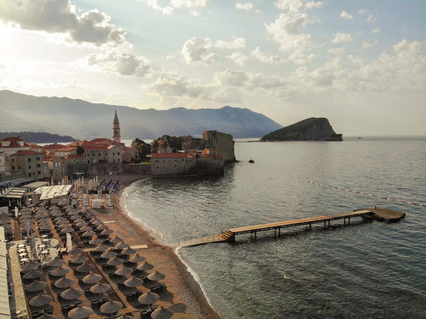 Avala Resort & Villas privātā pludmale ar saulessargiem un koka laipu Budvā — skats uz vecpilsētas mūriem un jūru