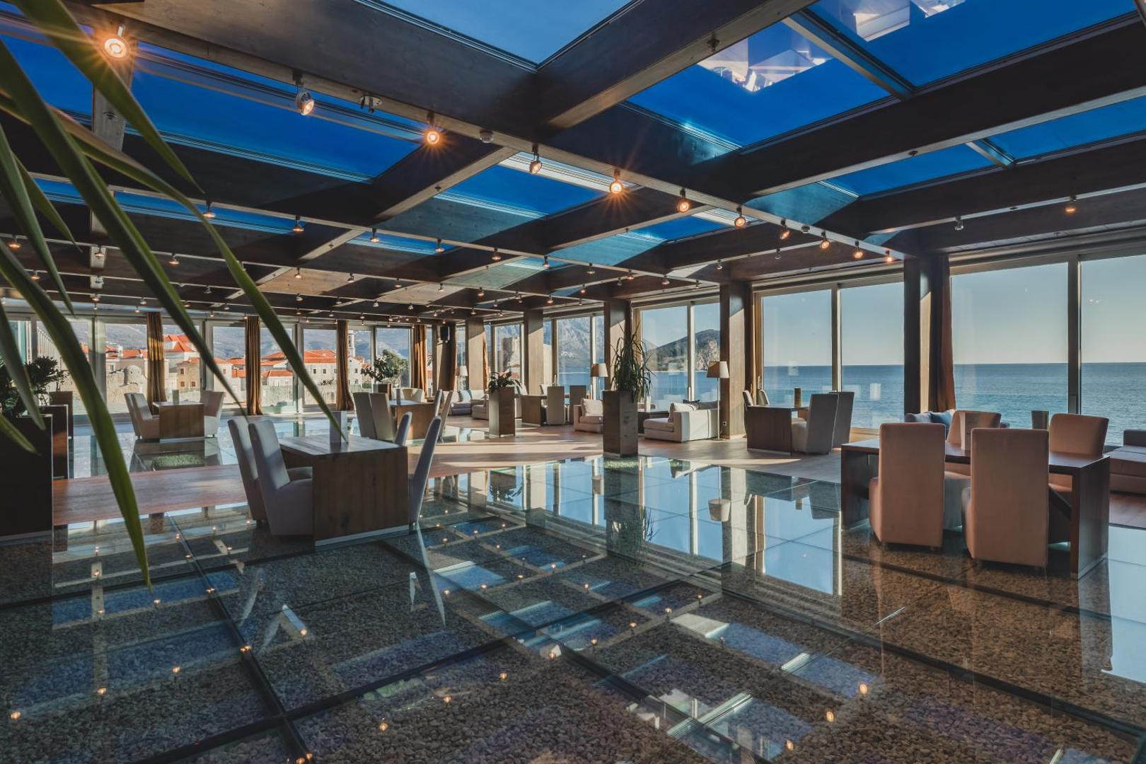 Avala Resort & Villas restorāns ar panorāmas logiem uz Adrijas jūru un Budvas vecpilsētu — gaišs interjers un stiklota grīda