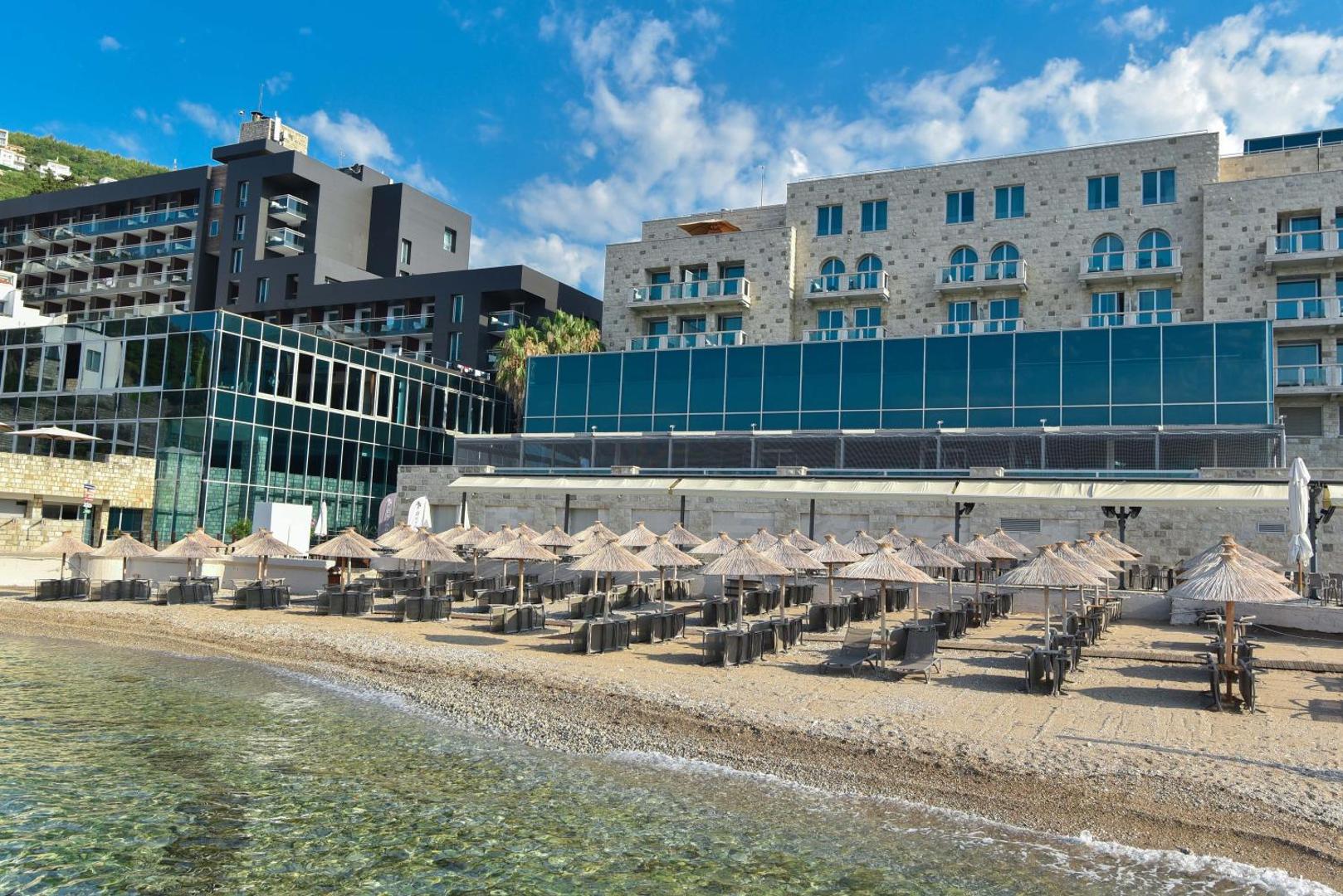 Avala Resort & Villas privātā oļu pludmale ar saulessargiem un zviļņiem Budvā — viesnīcas fasāde un dzidrs jūras ūdens