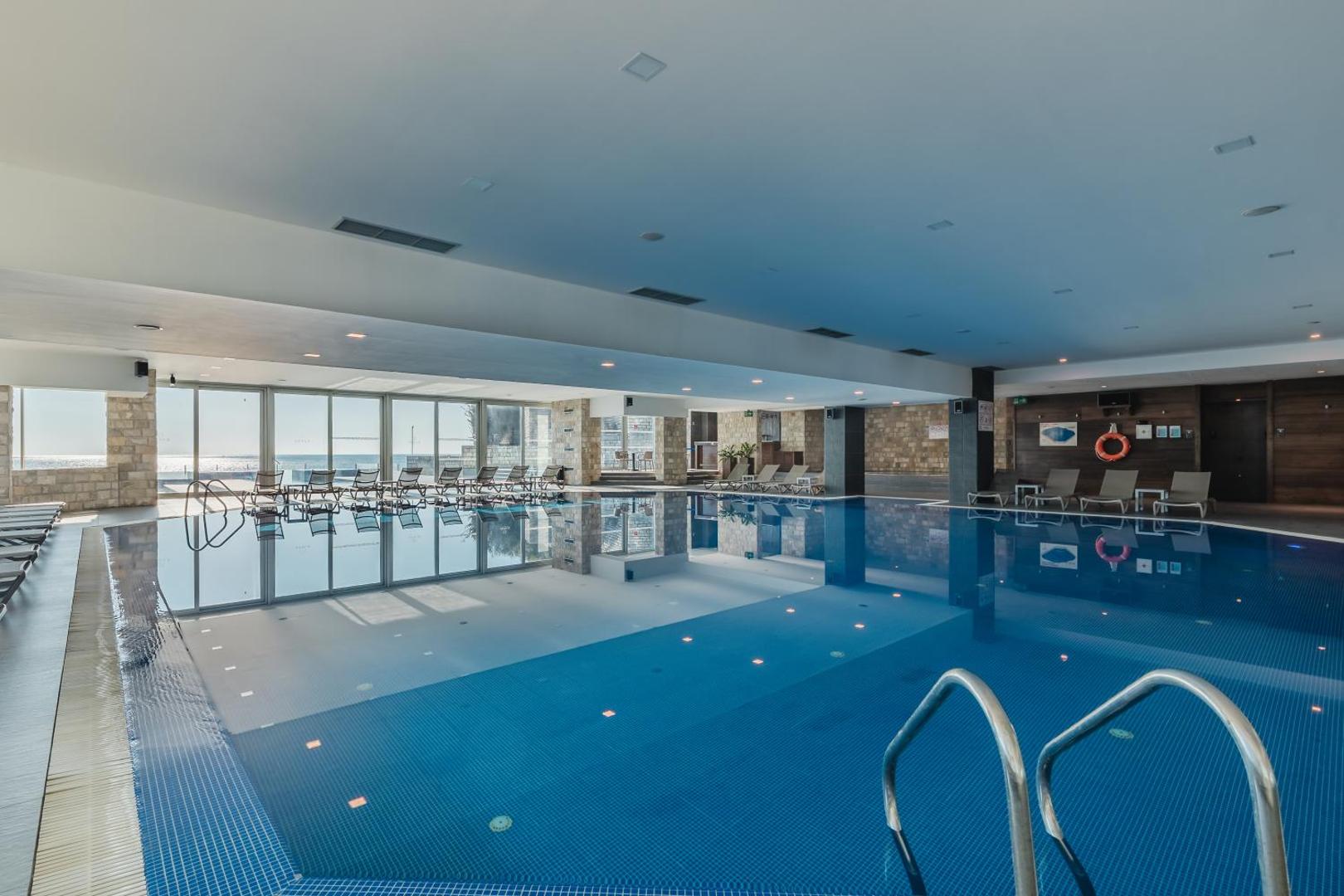 Avala Resort & Villas plašais iekštelpu baseins ar panorāmas logiem, sauļošanās krēsliem un skatu uz jūru