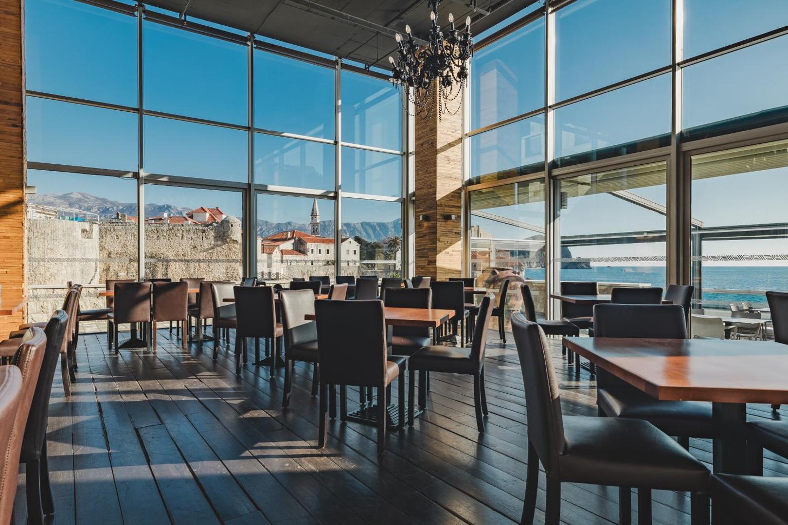 Avala Resort & Villas restorāna interjers ar koka grīdām un skatu uz Budvas vecpilsētas baznīcas torni un kalniem