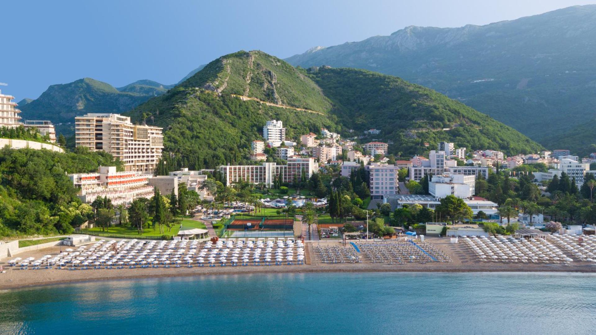 Iberostar Waves Bellevue privātā pludmale ar daudziem saulessargiem un gultām pie zilās Adrijas jūras uz kalnu fona Melnkalnē