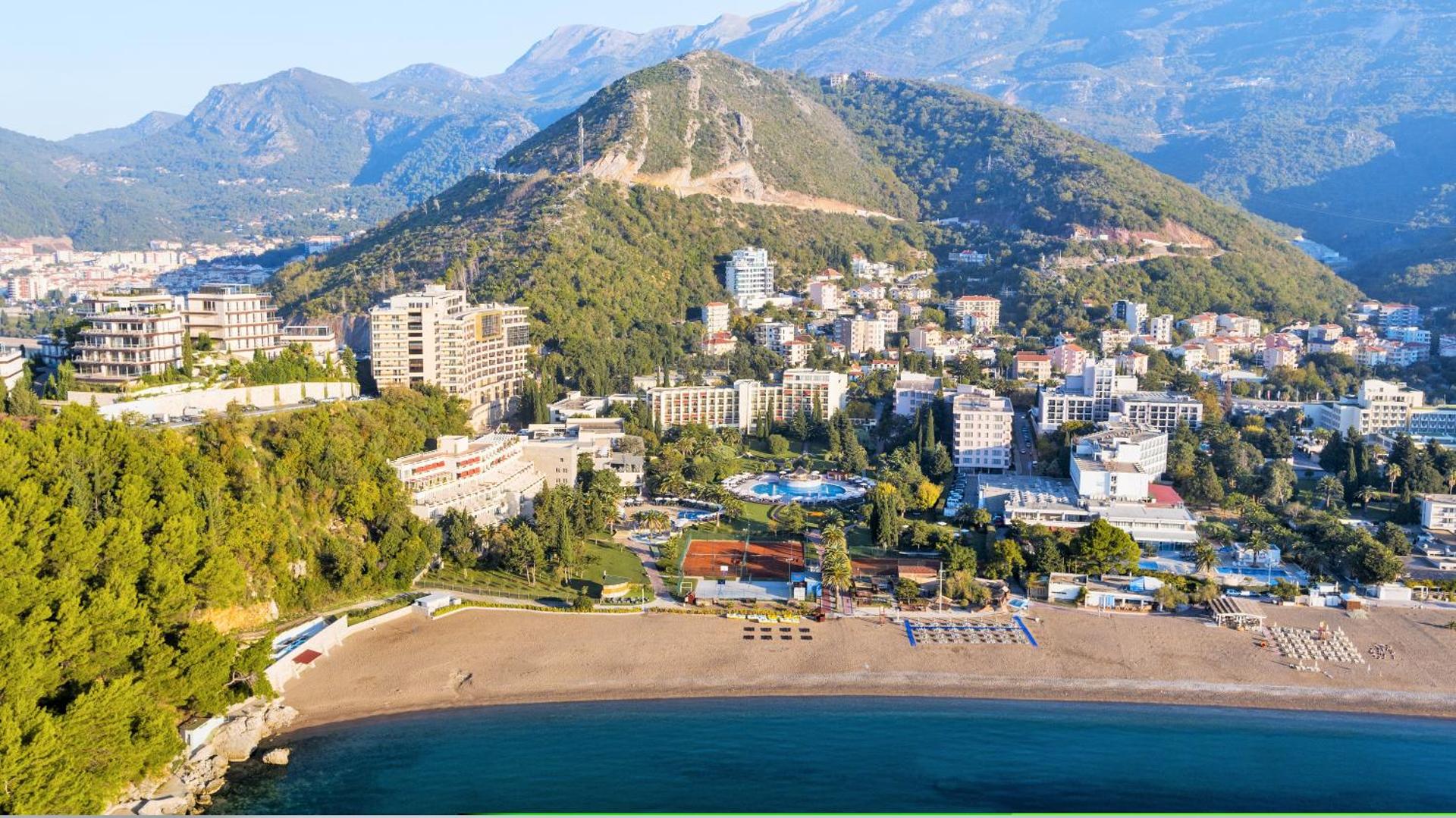 Iberostar Waves Bellevue panorāmas skats no putna lidojuma uz viesnīcas kompleksu, plašo pludmali un kalniem Bečiči kūrortā, Melnkalnē