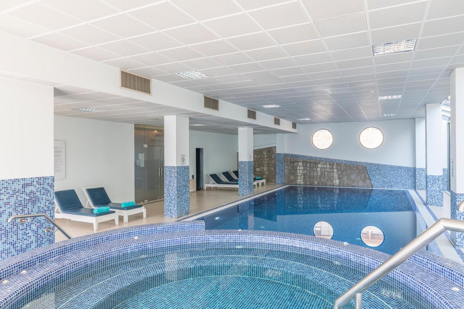 Iberostar Waves Bellevue viesnīcas iekštelpu baseins un džakuzi zona ar ziliem mozaīkas flīžu akcentiem un atpūtas krēsliem relaksācijai