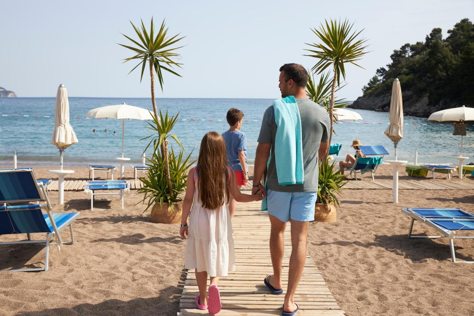 Ģimene dodas uz Iberostar Waves Bellevue privāto pludmali pa koka laipu gar saulessargiem un jūras krastu