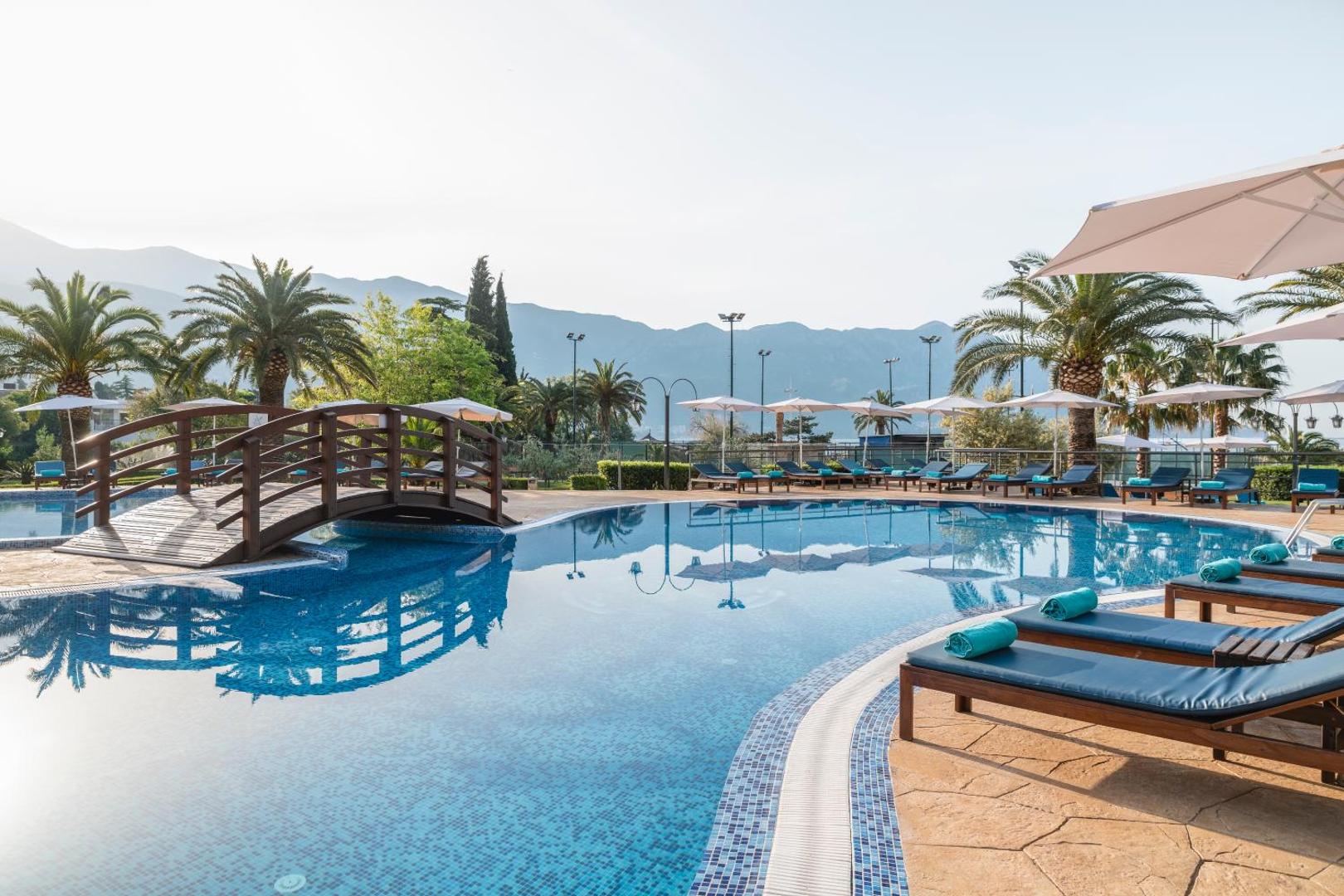 Iberostar Waves Bellevue āra baseins ar koka tiltiņu un sauļošanās krēsliem kalnu fonā Bečiči kūrortā, Melnkalnē