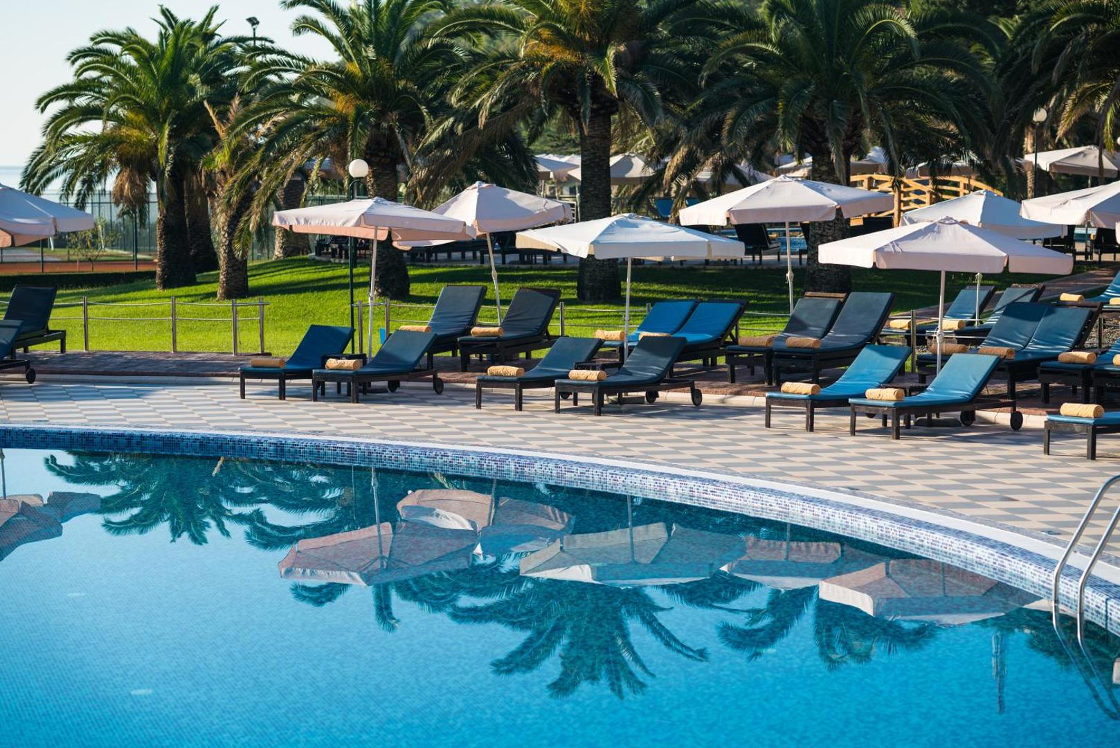 Viesnīcas Iberostar Waves Bellevue sauļošanās zona ar ziliem krēsliem un saulessargiem palmu ielenkumā pie baseina