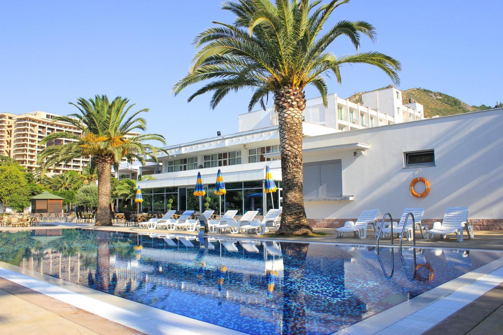 Montenegro Beach Resort galvenais āra baseins pie viesnīcas ēkas ar atpūtas krēsliem un eksotiskām palmām saulainā dienā