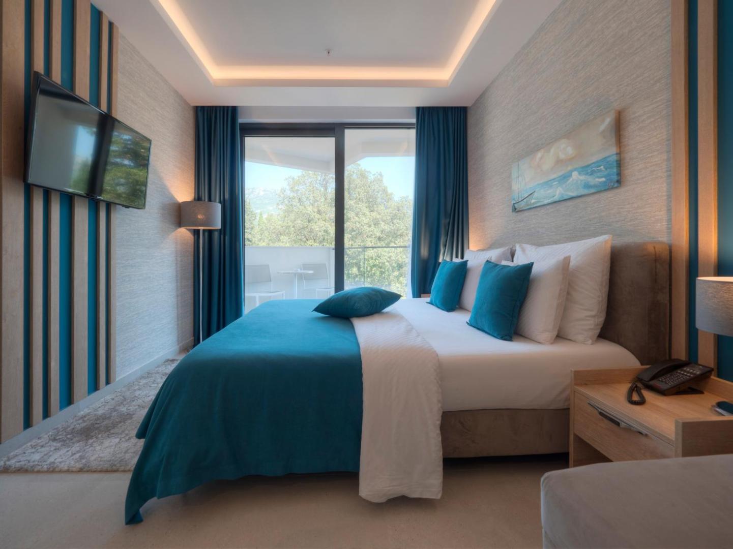 Montenegro Beach Resort guļamistaba ar lielu gultu, tirkīza akcentiem un skatu caur logu uz zaļojošu apkārtni