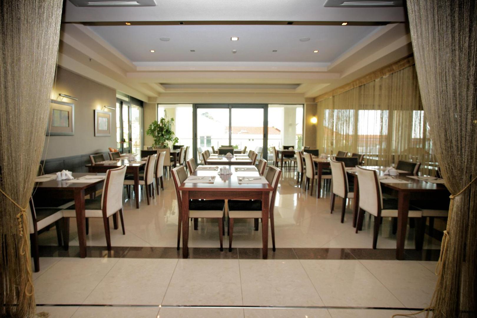 Monte Casa Spa & Wellness elegantā restorāna zāle ar klātiem galdiem, lieliem logiem un modernu interjeru