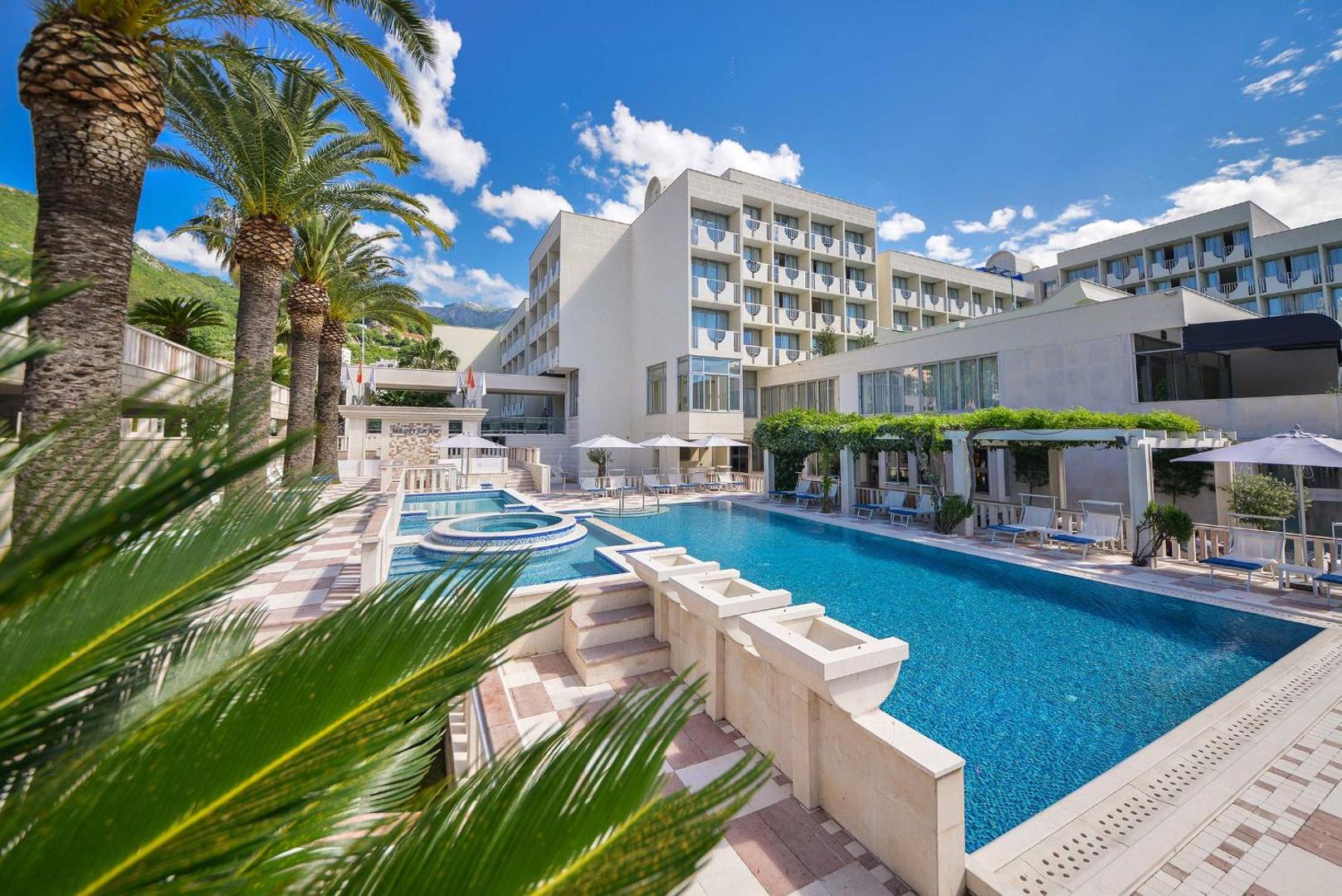 Hotel Mediteran āra baseins Bečiči kūrortā Melnkalnē — skats uz viesnīcas ēku, palmām un sauļošanās terasi ar saulessargiem