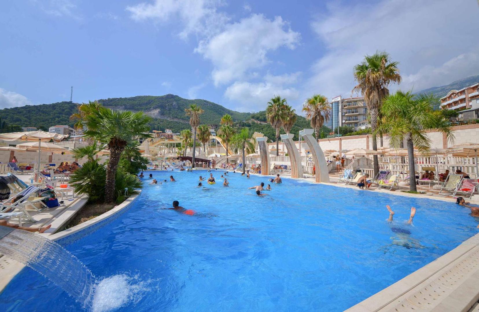 Lielais atpūtas baseins Hotel Mediteran teritorijā ar ūdens kaskādēm un atpūtniekiem — fonā kalnu ainava un pilsētas apbūve