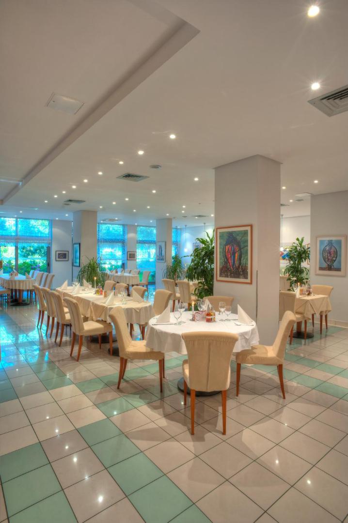 Hotel Rivijera iekštelpu restorāns ar gaišu interjeru, apaļiem galdiem, baltiem galdautiem un modernu flīžu grīdu