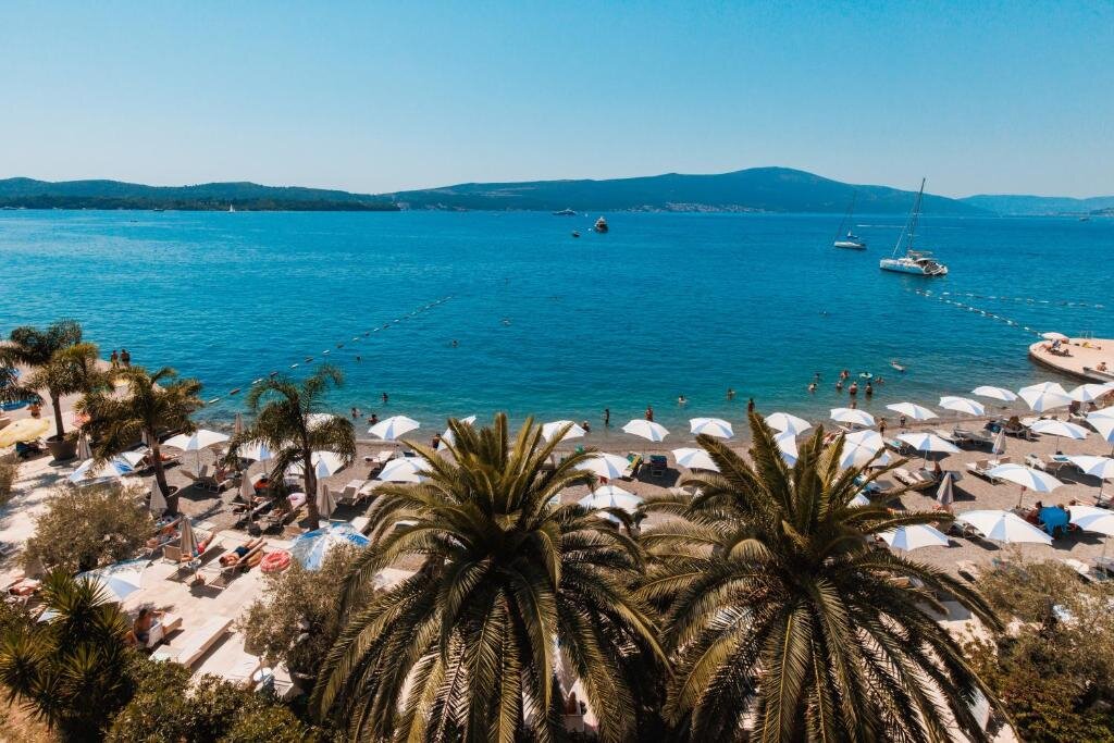 Viesnīcas Palma pludmale ar daudziem saulessargiem, palmām un atpūtniekiem pie zila līča ūdens saulainā dienā