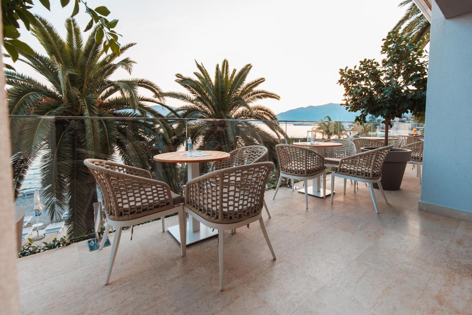 Hotel Palma āra terase ar pītām mēbelēm un palmām Tivatas piekrastē, Melnkalnē — skats uz Adrijas jūru un kalniem saulrietā