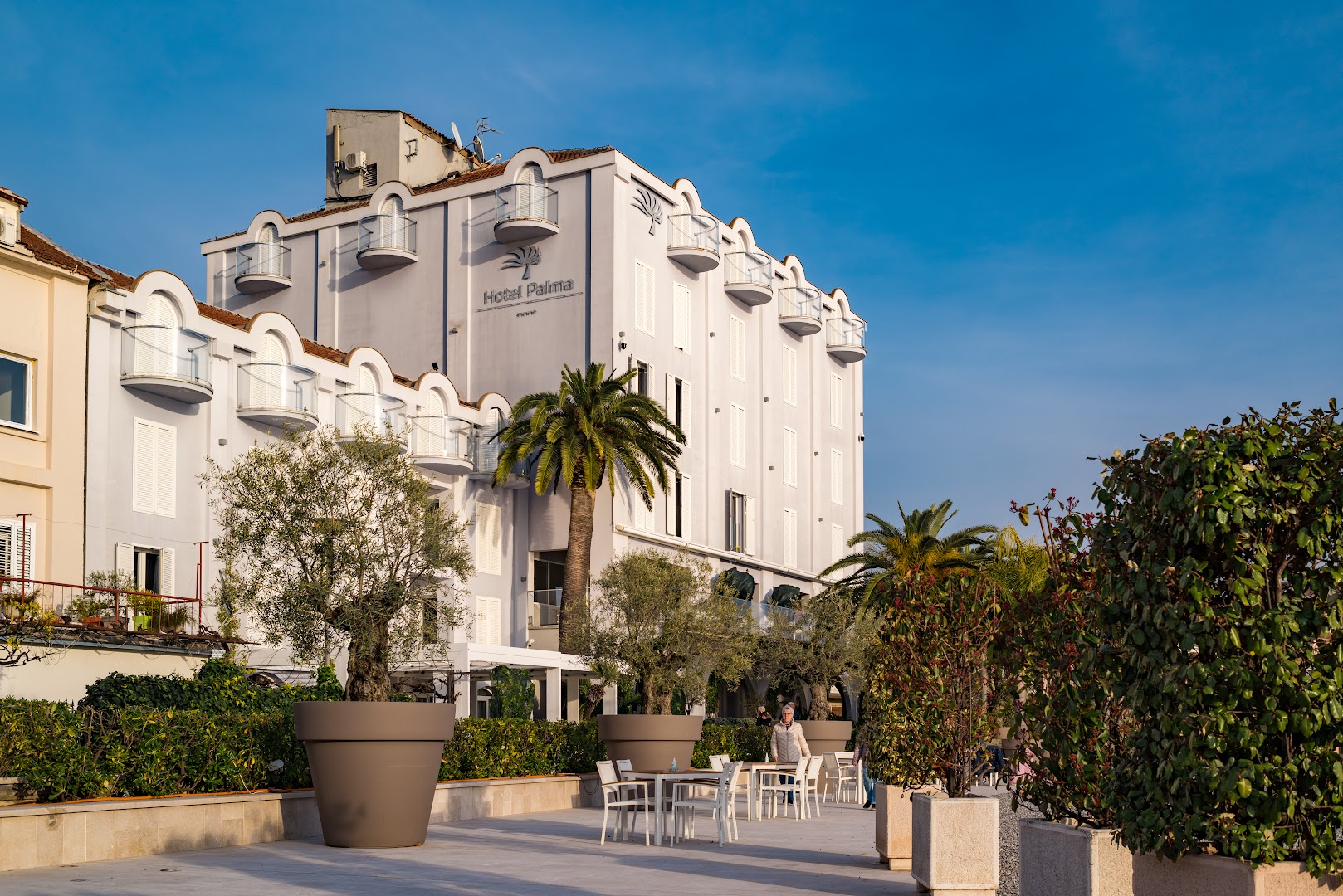 Hotel Palma galvenā ēka un āra terase ar galdiņiem Tivatas kūrortā — gaiša arhitektūra, palmas un dekoratīvie augi podos