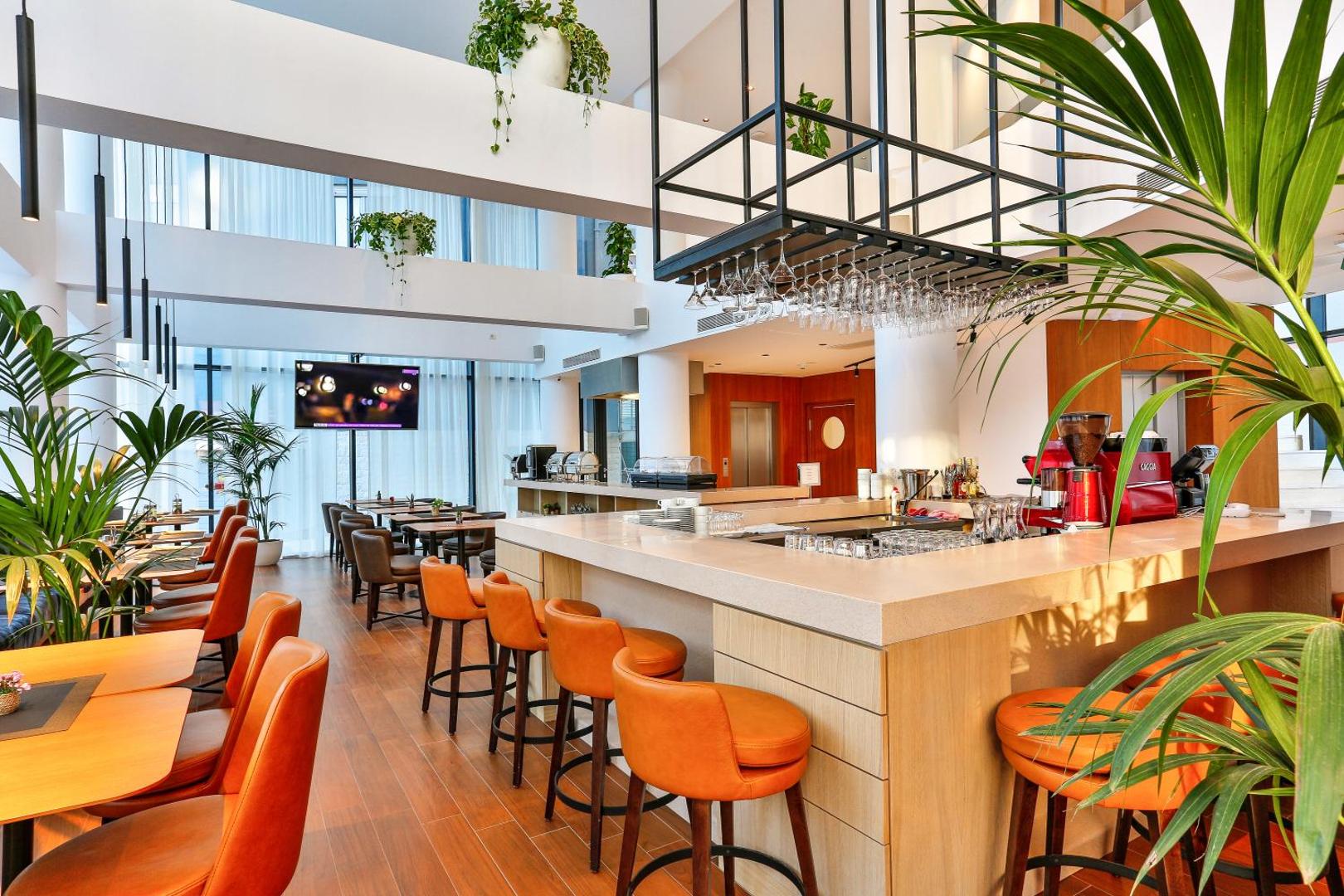 Hotel Fagus by Aycon modernais bārs un restorāns ar oranžiem krēsliem, augstiem griestiem un zaļiem telpaugiem