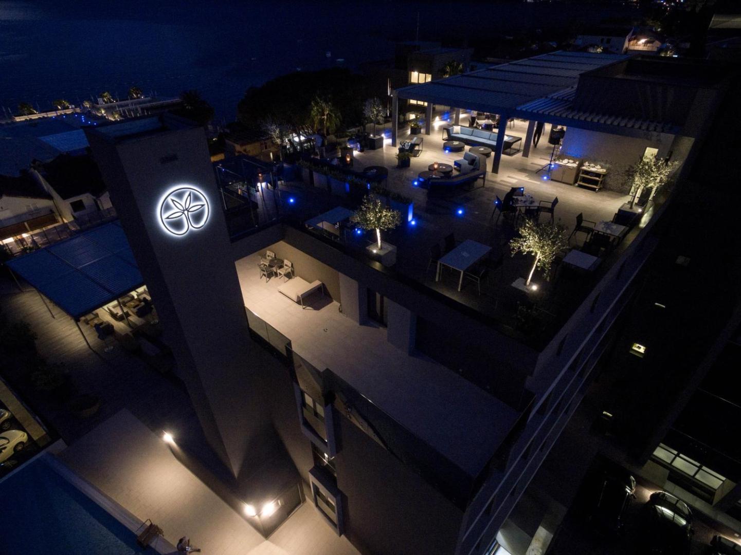 Palmon Bay Hotel & SPA jumta terase naktī ar izgaismotu logo, galdiņiem un skatu uz jūru Melnkalnē