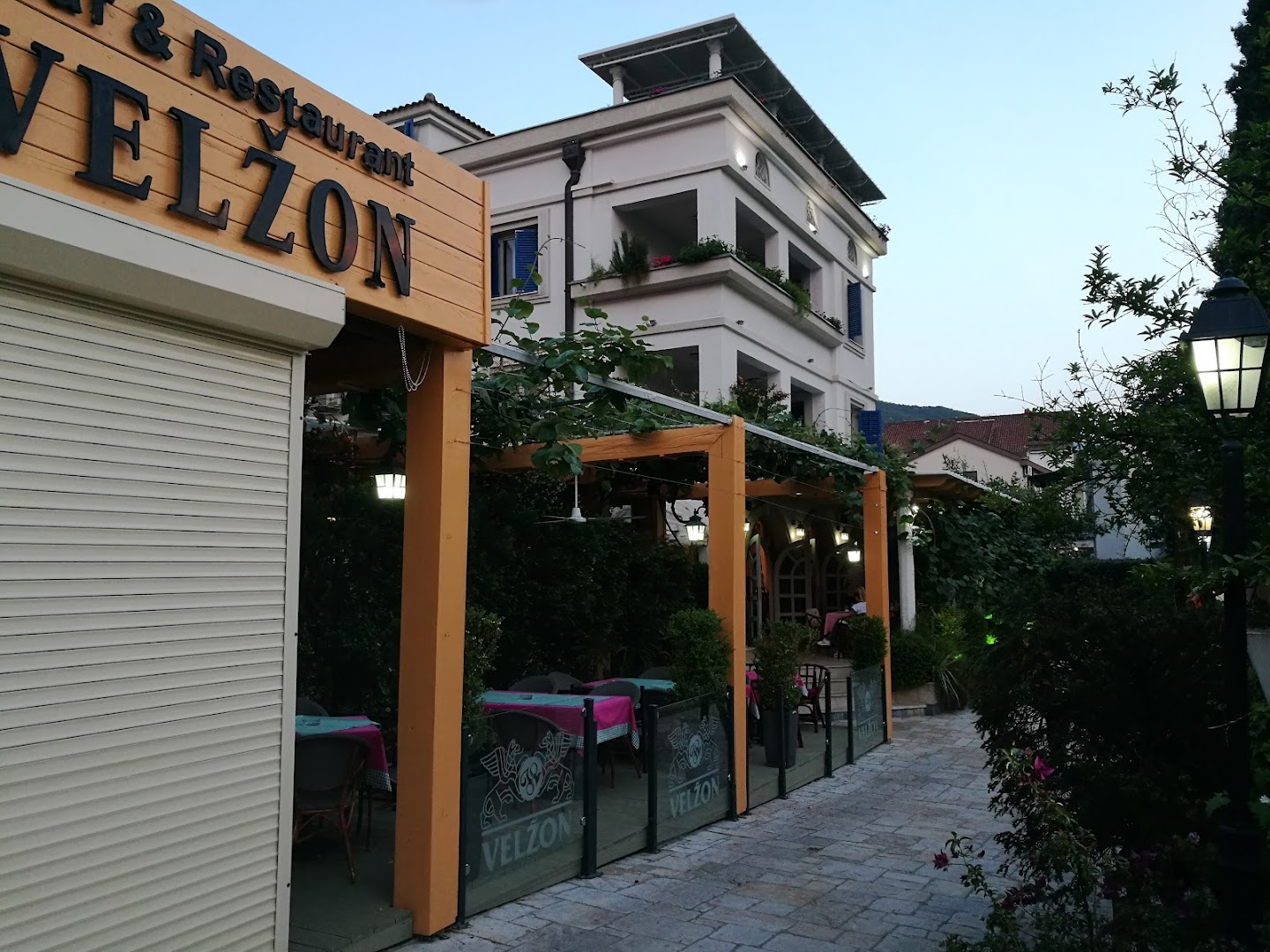 Villa Velzon viesnīcas ēka un restorāna ieeja vakarā Budvā — izgaismota fasāde un mājīga terase pie ielas