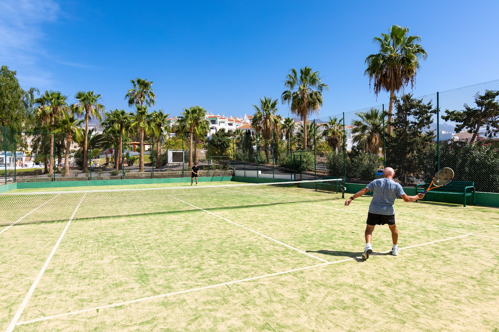 Tropical Park tenisa korts Tenerifē — viesi spēlē tenisu saulainā dienā palmu un viesnīcas ēku fonā