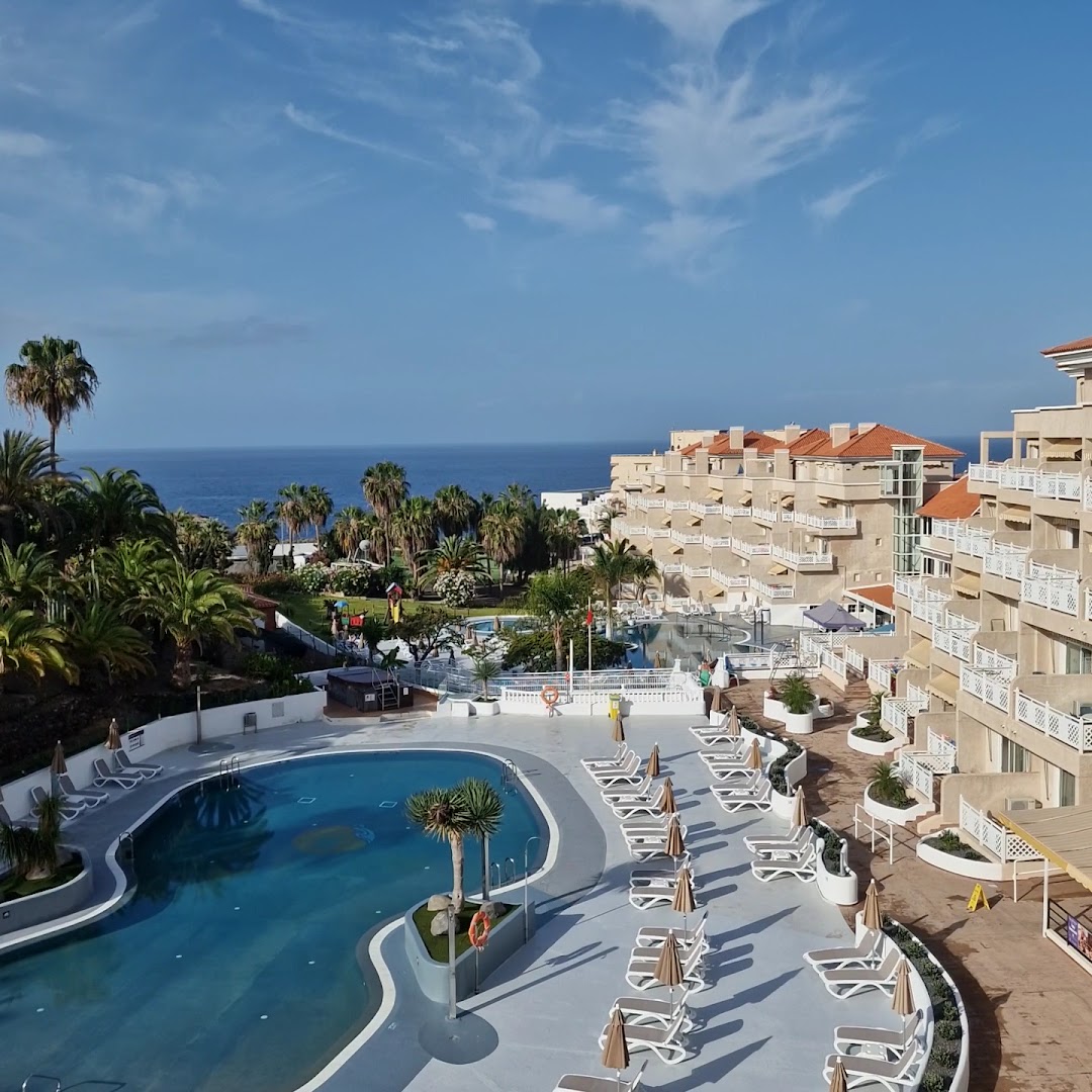 Tropical Park viesnīcas galvenais baseins ar atpūtas krēsliem un skatu uz okeānu Tenerifes piekrastē, ieskauts palmās un apartamentos