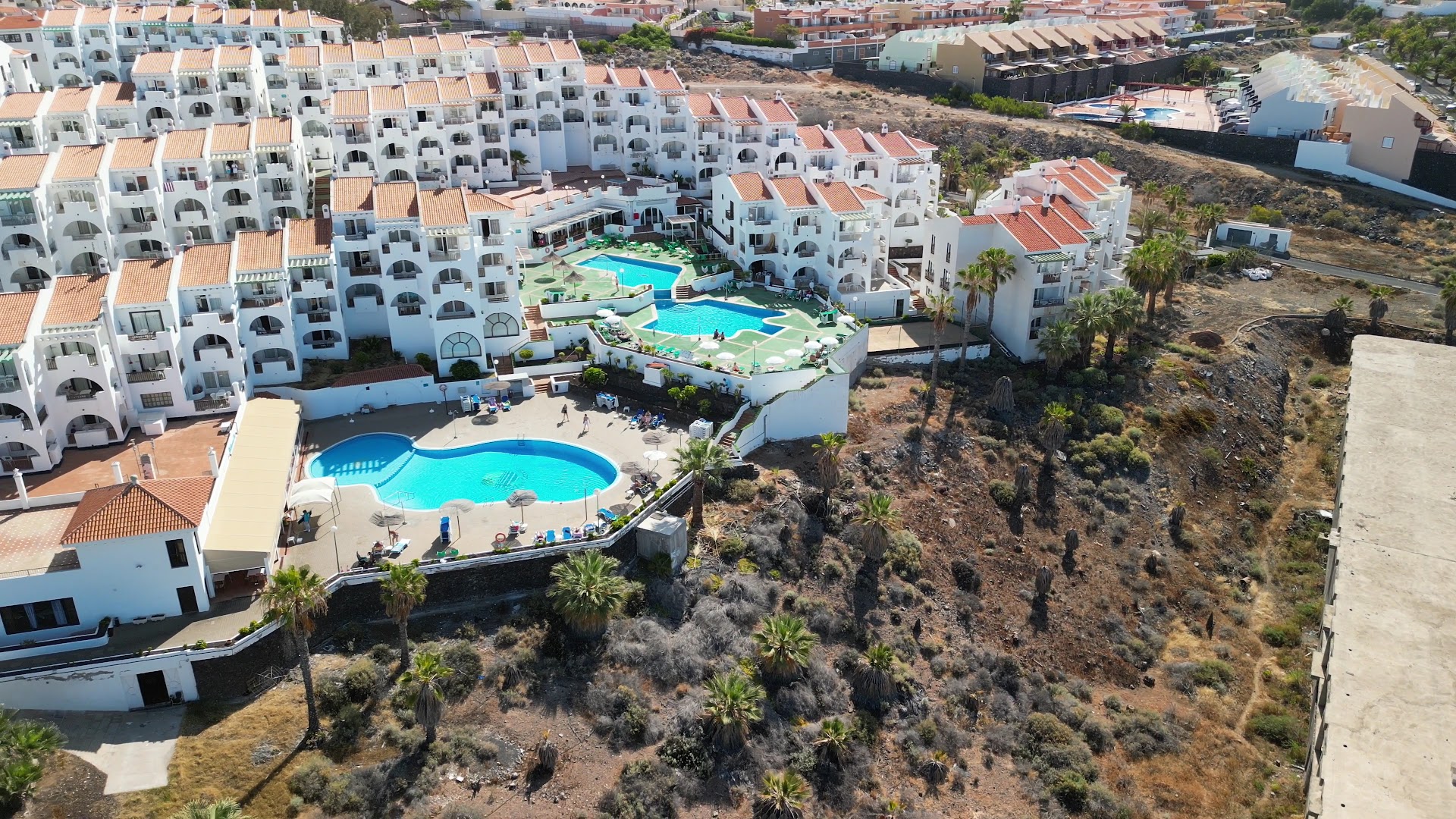 Aparthotel Tropical Park kompleksa skats no putna lidojuma Tenerifē — balti korpusi ar vairākiem āra baseiniem un sauļošanās terasēm piekrastē
