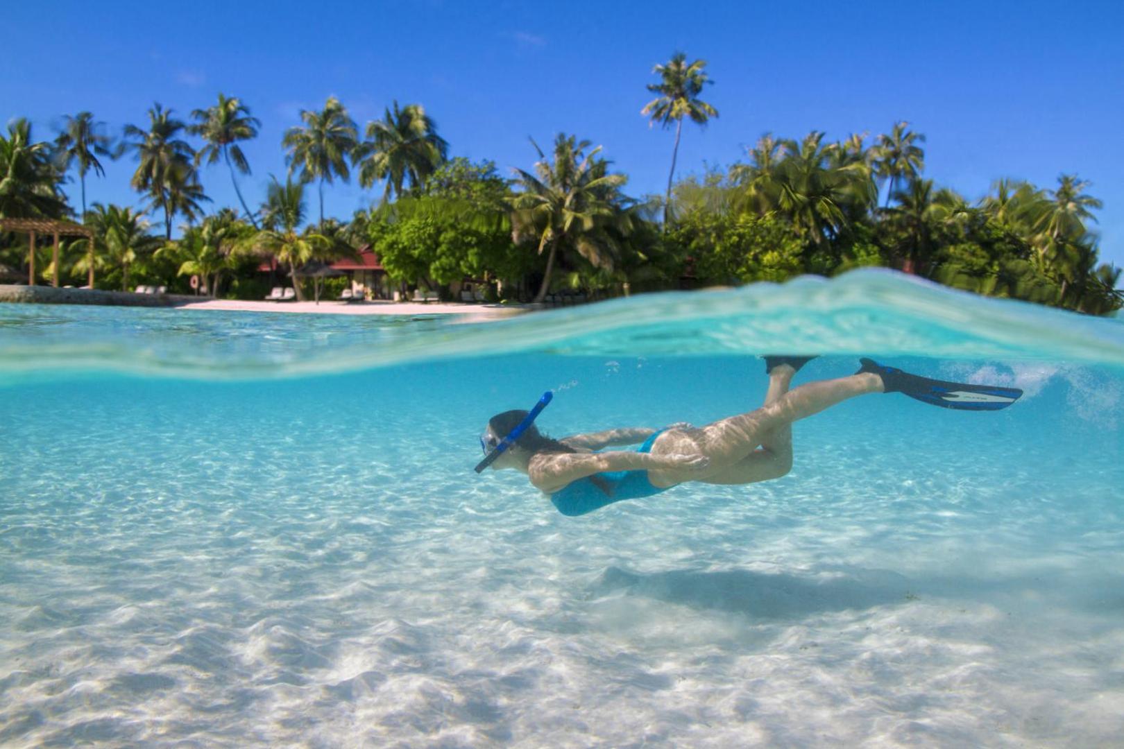 Sieviete peld zem ūdens Kurumba Maldives piekrastē, fonā redzama balto smilšu pludmale un tropiskas palmas