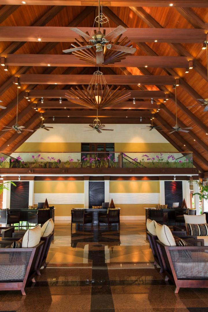 Kurumba Maldives viesnīcas lobby zona ar augstiem koka griestiem, masīvām lustrām un ērtiem atpūtas krēsliem