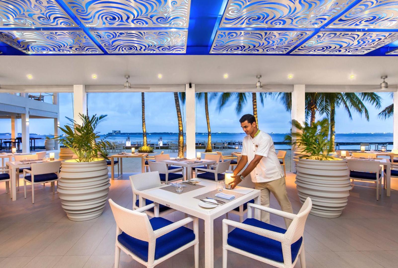 Kurumba Maldives restorāns ar skatu uz okeānu — viesmīlis klāj galdus modernā terasē ar ziliem akcentiem un dekoratīviem griestiem