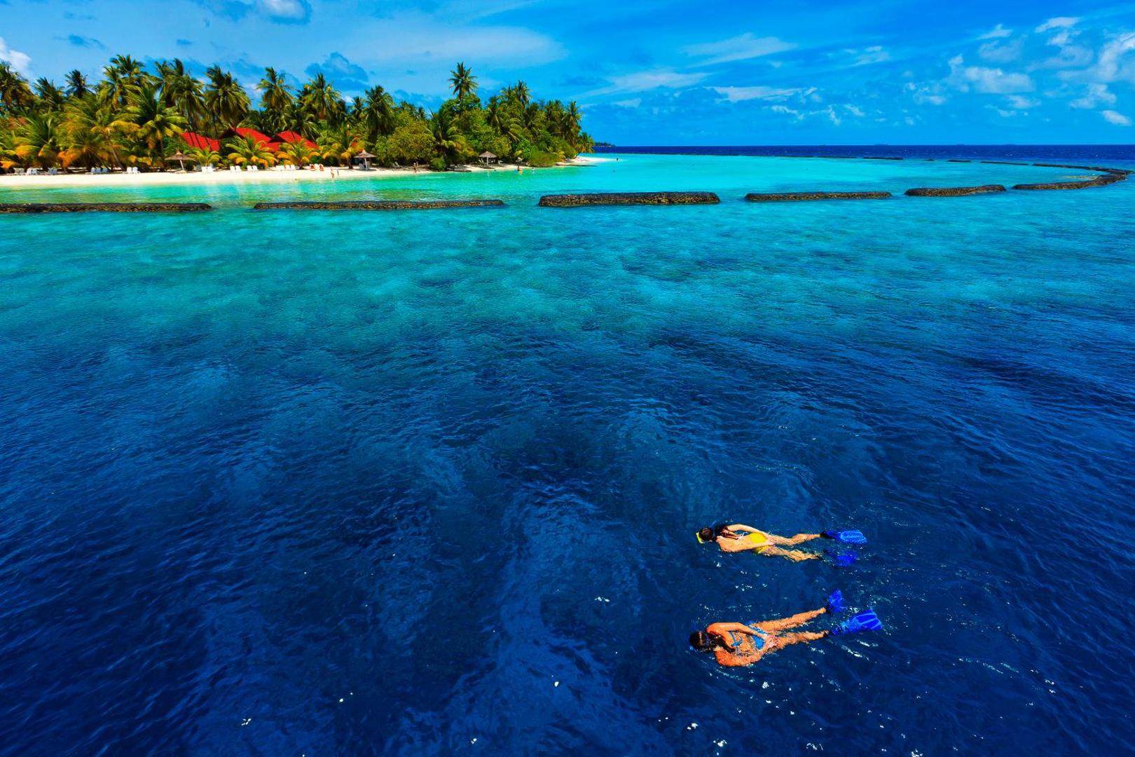 Aerofoto skats uz Kurumba Maldives salu ar palmu birzi, baltām smiltīm un diviem tūristiem, kuri peld dzidrā okeāna ūdenī