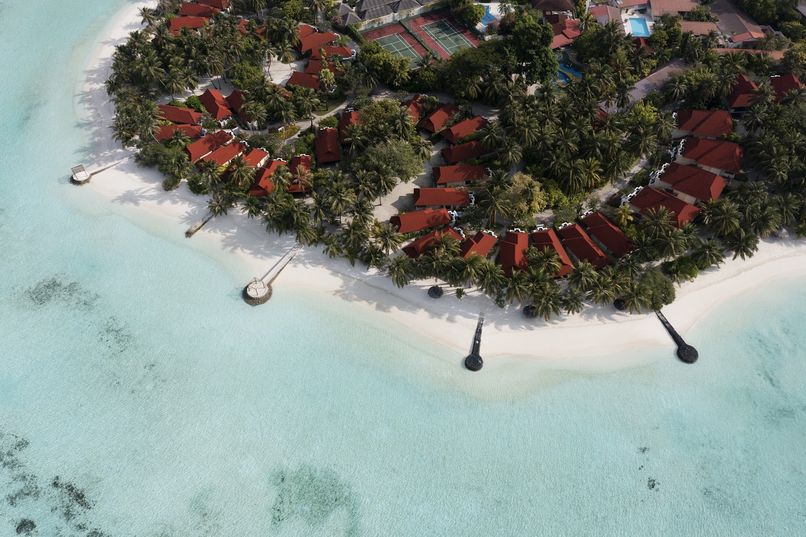 Kurumba Maldives bungalo komplekss ar sarkaniem jumtiem starp palmām un tenisa kortiem pie tirkīzzila okeāna krasta.