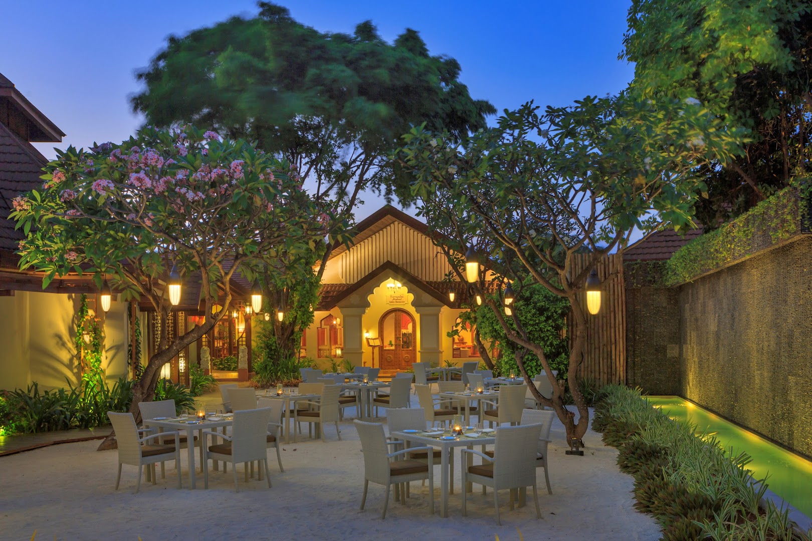 Niva Kurumba Maldives restorāna āra terase vakarā ar balti klātiem galdiem smiltīs un izgaismotiem kokiem