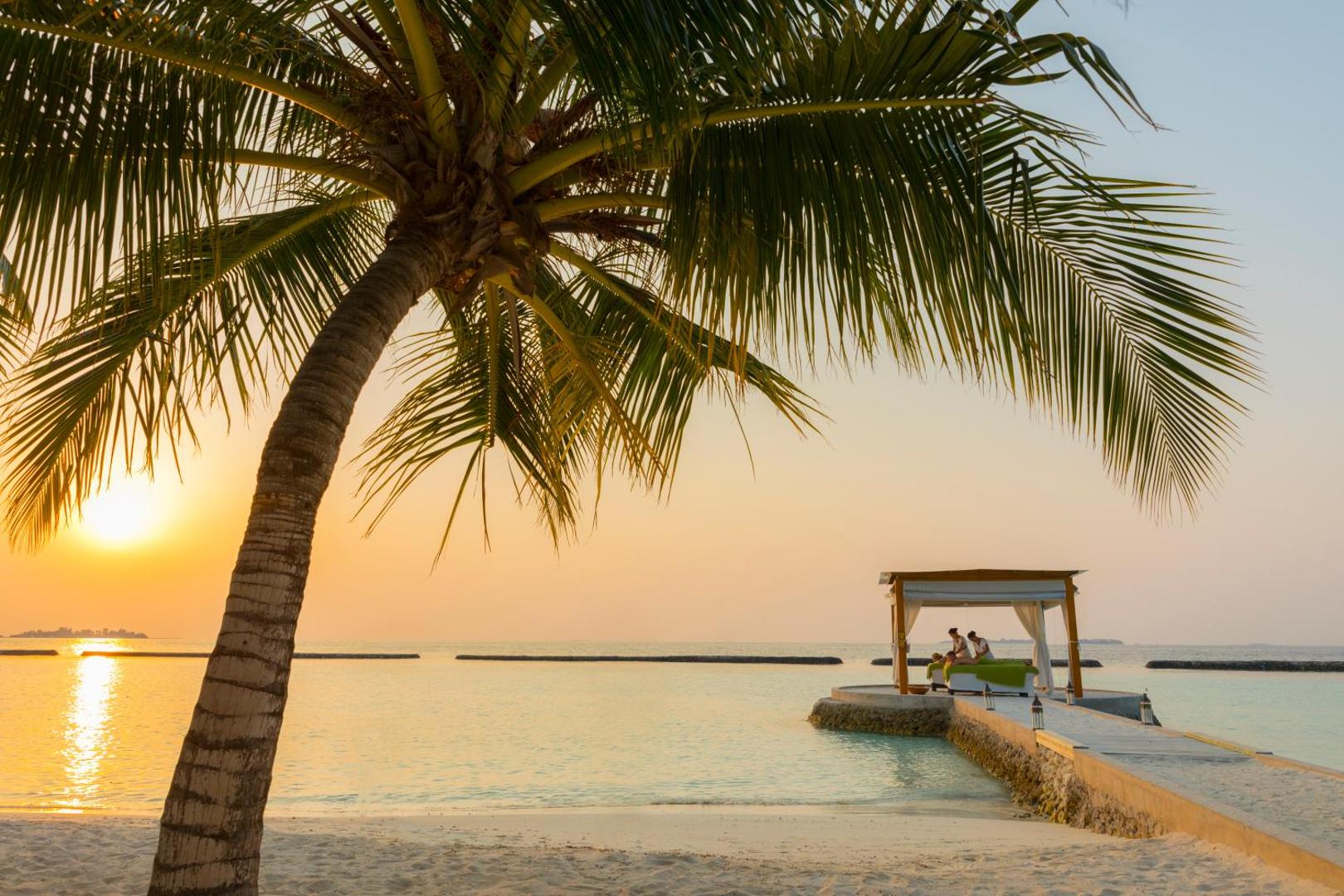 Kurumba Maldives saulrieta skats no pludmales — palma priekšplānā un spa paviljons uz mola okeāna piekrastē