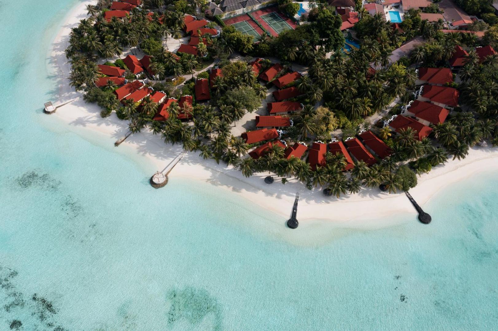 Kurumba Maldives aerofoto ar pludmales joslu, bungalo rindu un dzidru lagūnas ūdeni saulainā dienā.